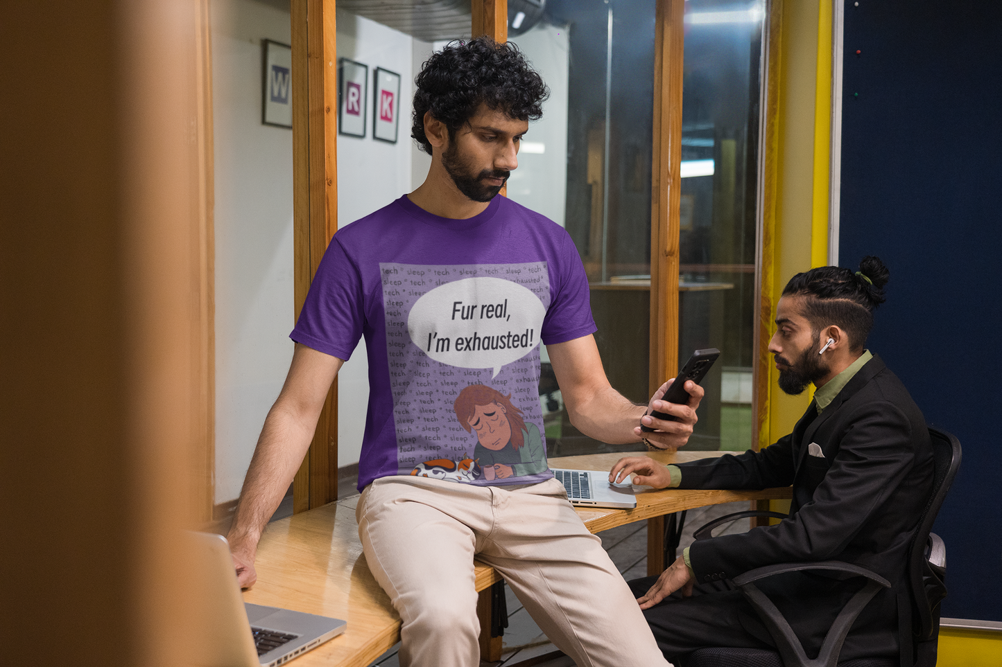 Man with curly hair and beard wearing a purple Fur Real, I'm Exhausted t-shirt while working in an office