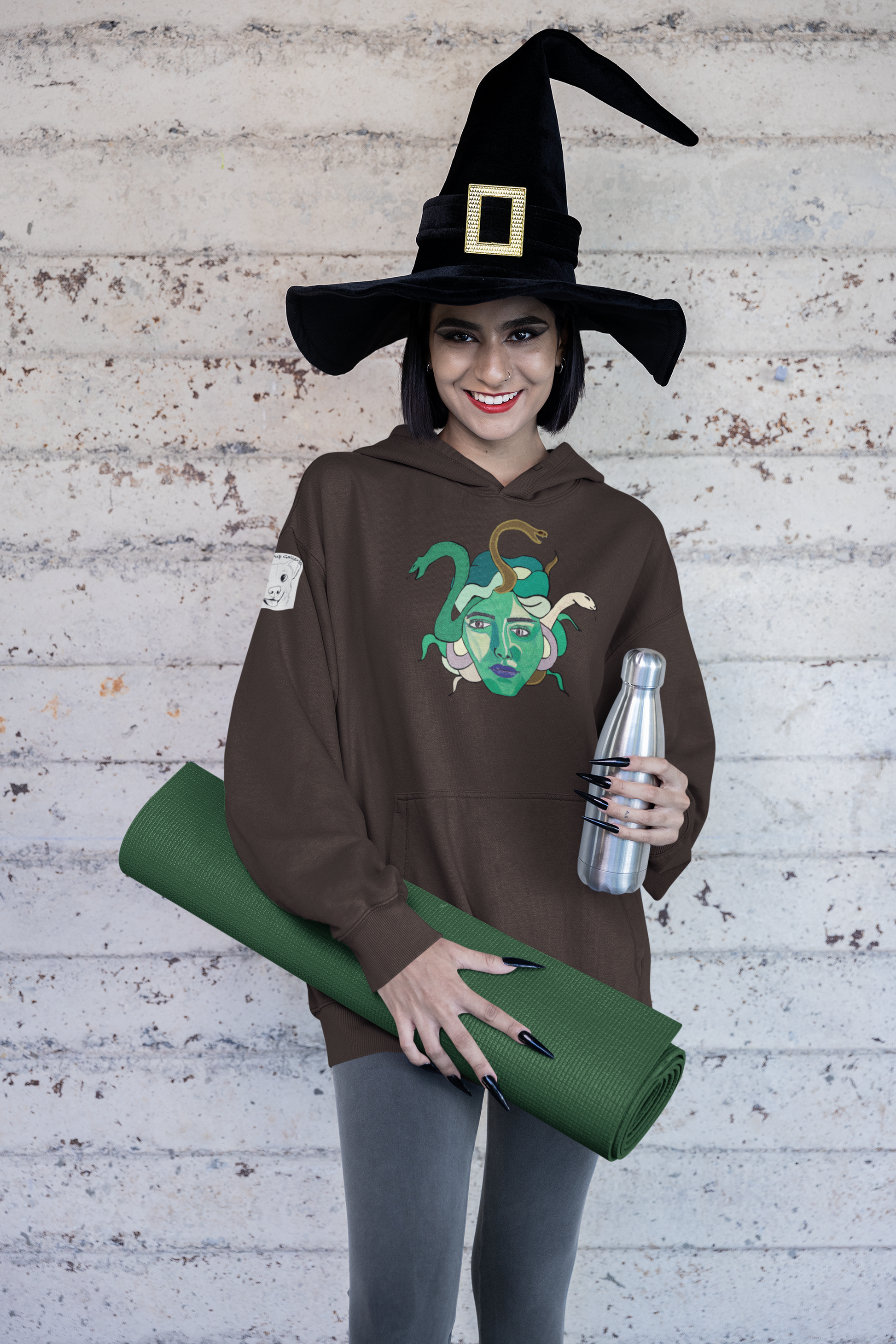 Woman in a witch hat holding a yoga mat and water bottle, wearing a dark chocolate Medusa hoodie by GrungePup Gallery