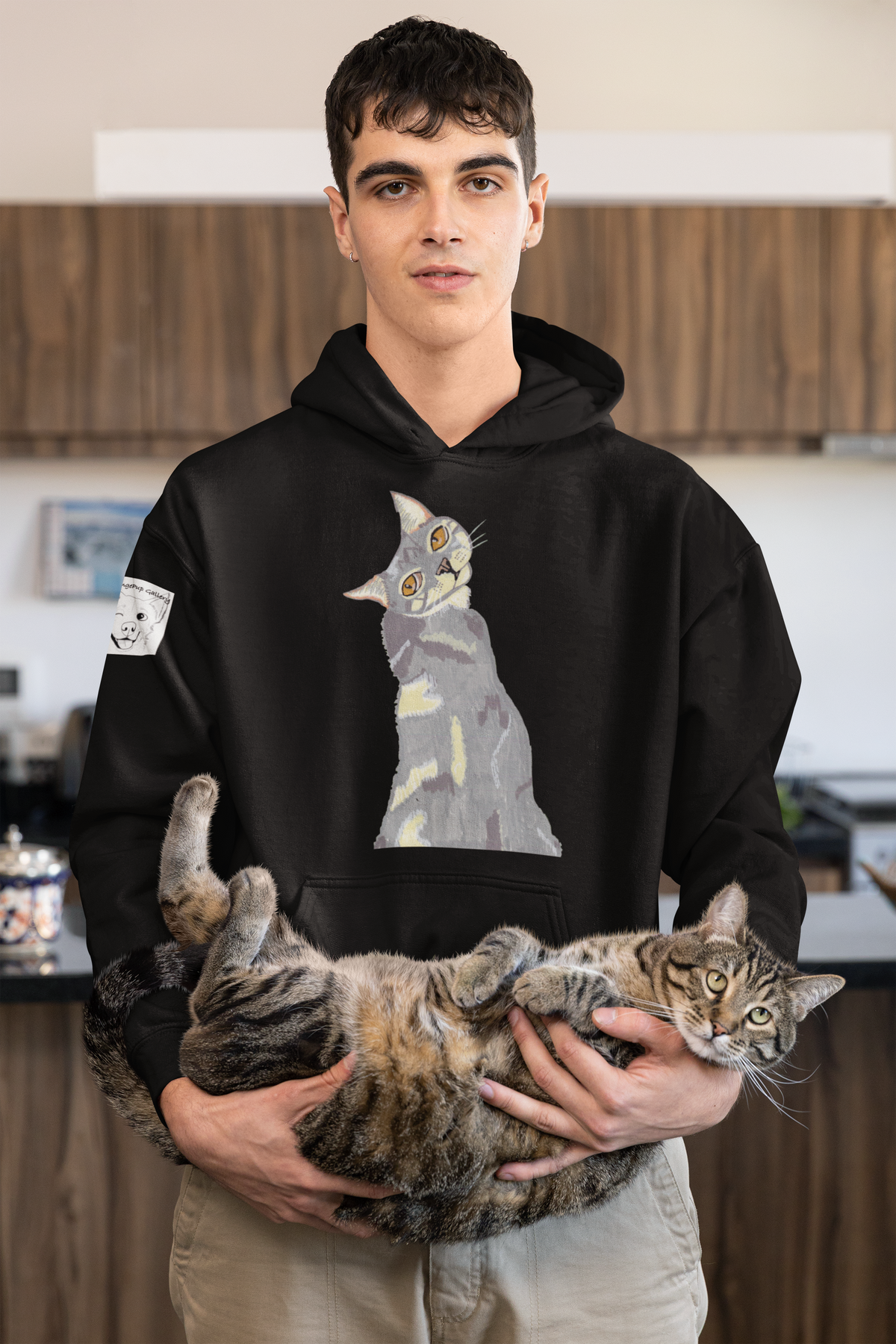 Man in a kitchen holding a tabby cat, wearing the Mr. Grey Cat Portrait Hoodie in black.