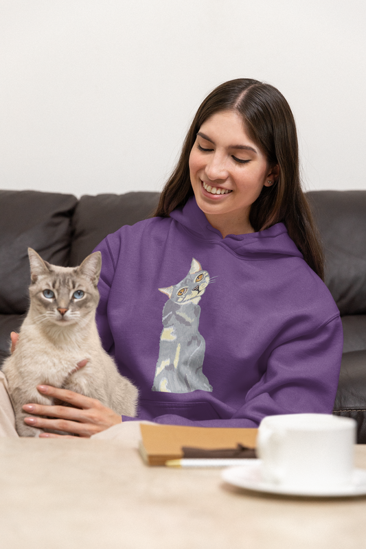 Smiling woman on a couch holding a light-colored cat, wearing the Mr. Grey Cat Portrait Hoodie in purple.