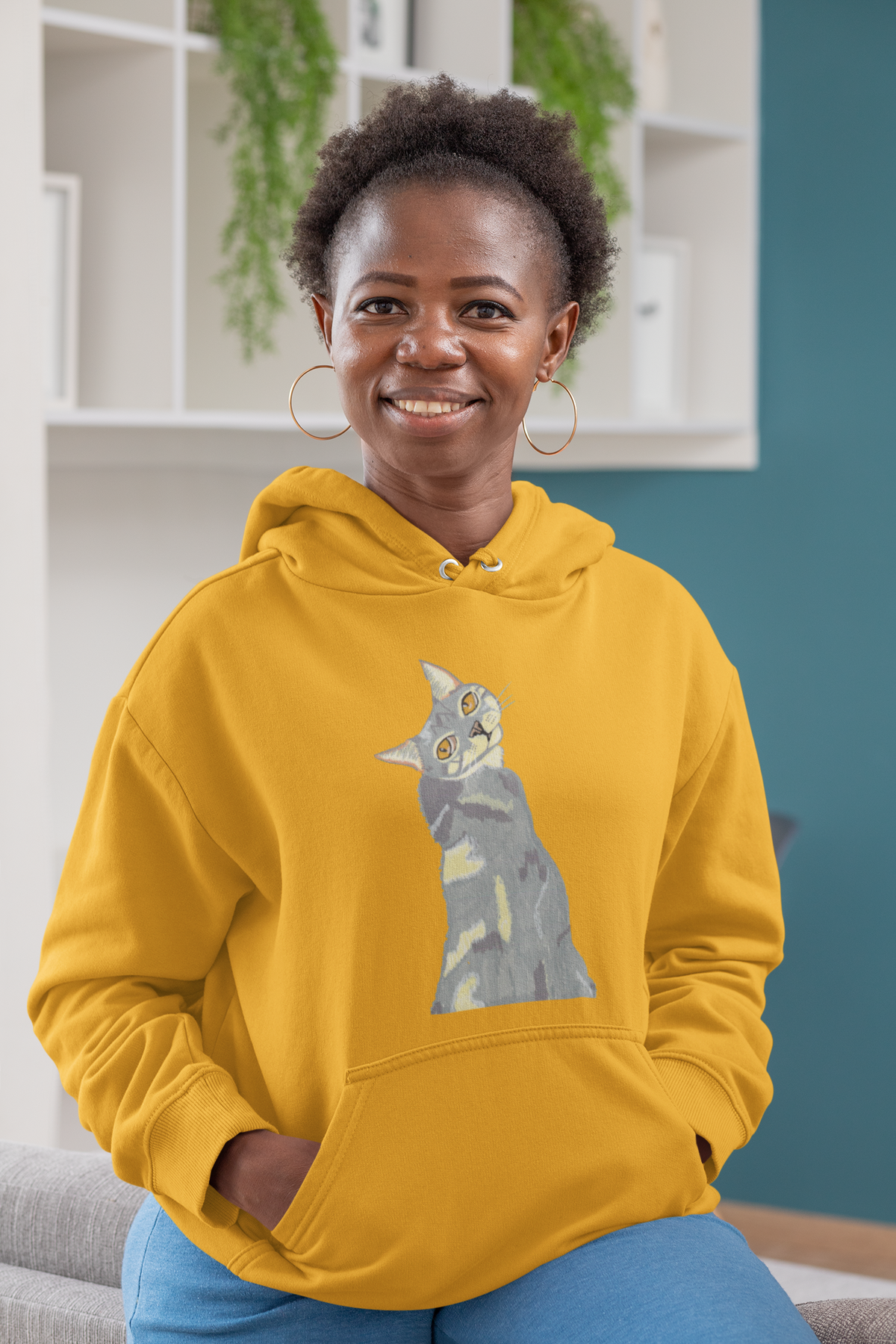 Smiling woman with short hair sitting indoors, wearing the Mr. Grey Cat Portrait Hoodie in team gold.