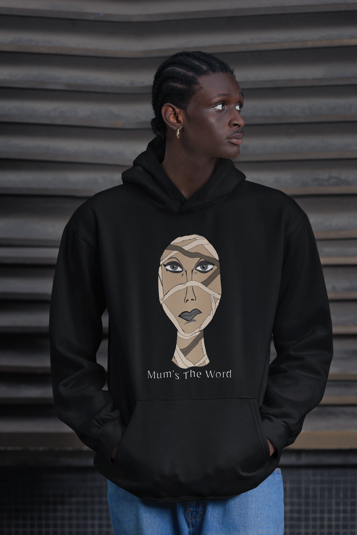 Man with cornrows standing in front of a metal wall wearing a black hoodie with the wrapped mummy face and Mum’s The Word text