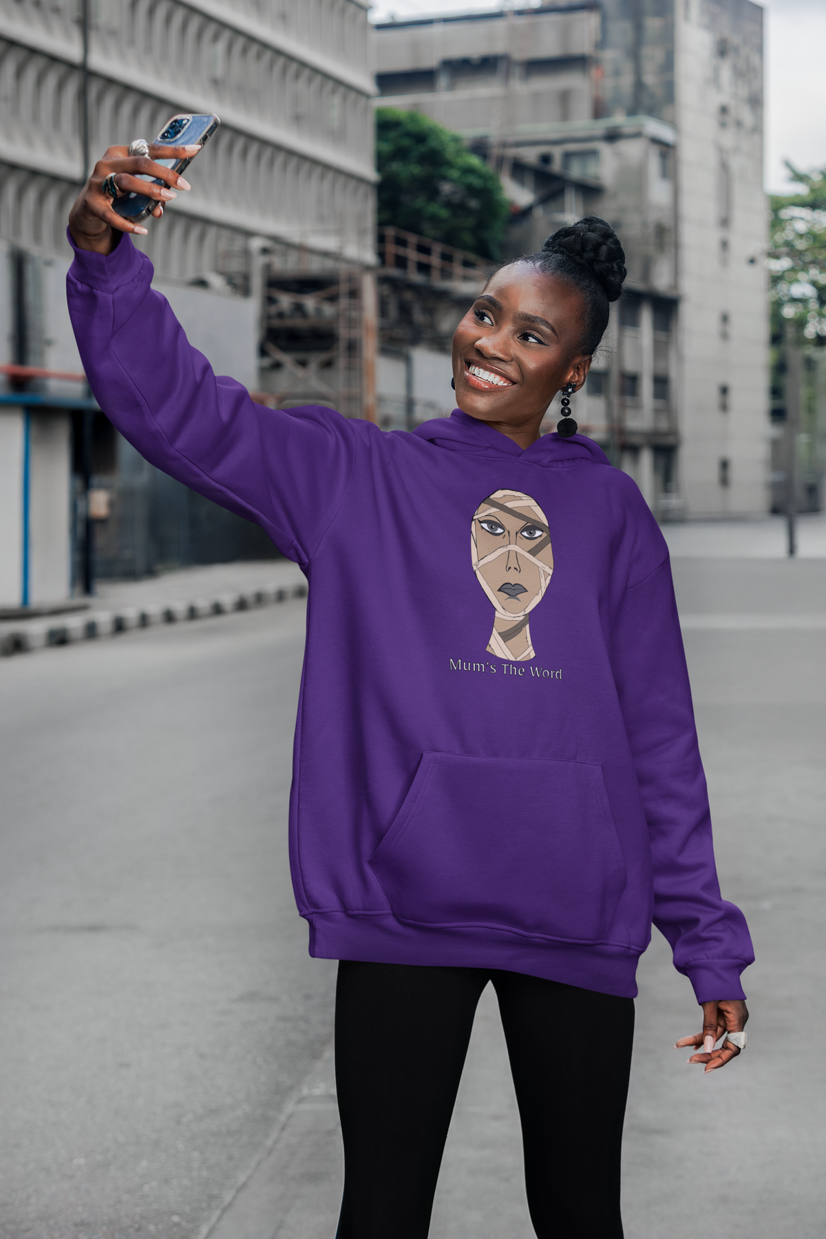 Woman taking a selfie on a city sidewalk in a purple hoodie with the mummy face and Mum’s The Word design