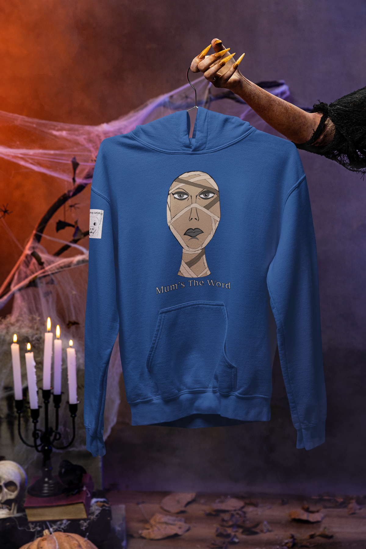 Royal blue Gildan hoodie on a hanger held by a clawed monster hand in a haunted studio, mummy face and Mum’s The Word print on the chest