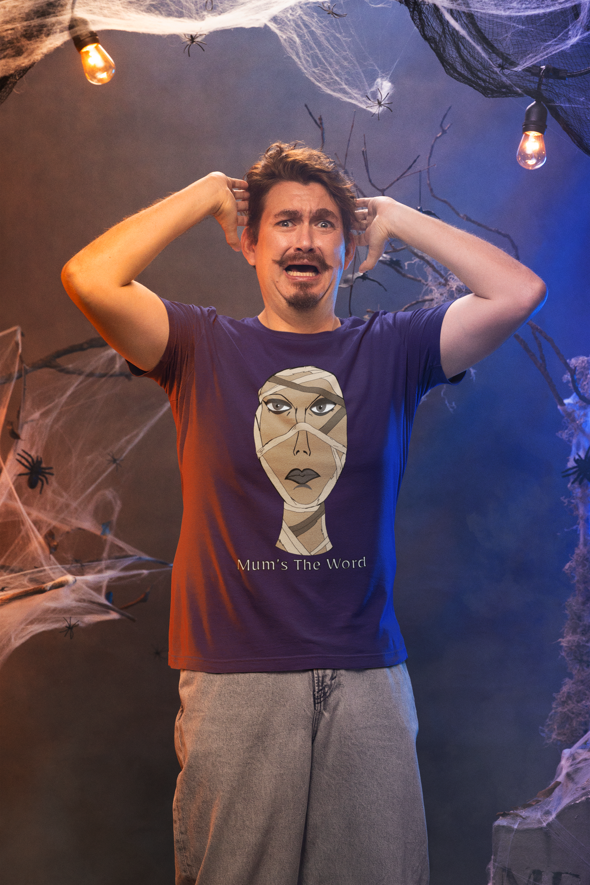 Scared man with hands on his head in a haunted house wearing a team purple t-shirt with the mummy face and Mum’s The Word design