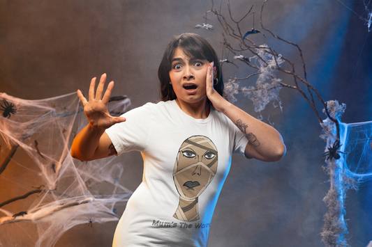 Woman with a scared expression in a white t-shirt reaching forward in a haunted house set, mummy face and Mum’s The Word text