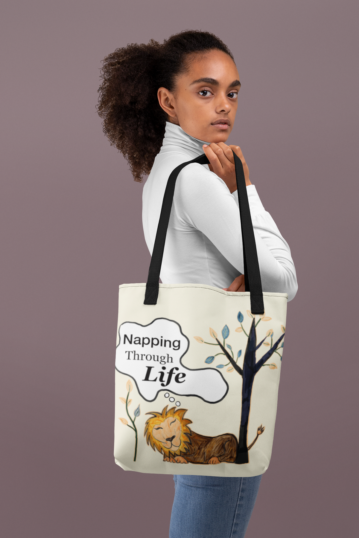Cream tote with black straps featuring the Napping Through Life design. Model in white turtleneck holding the bag against a solid mauve background.
