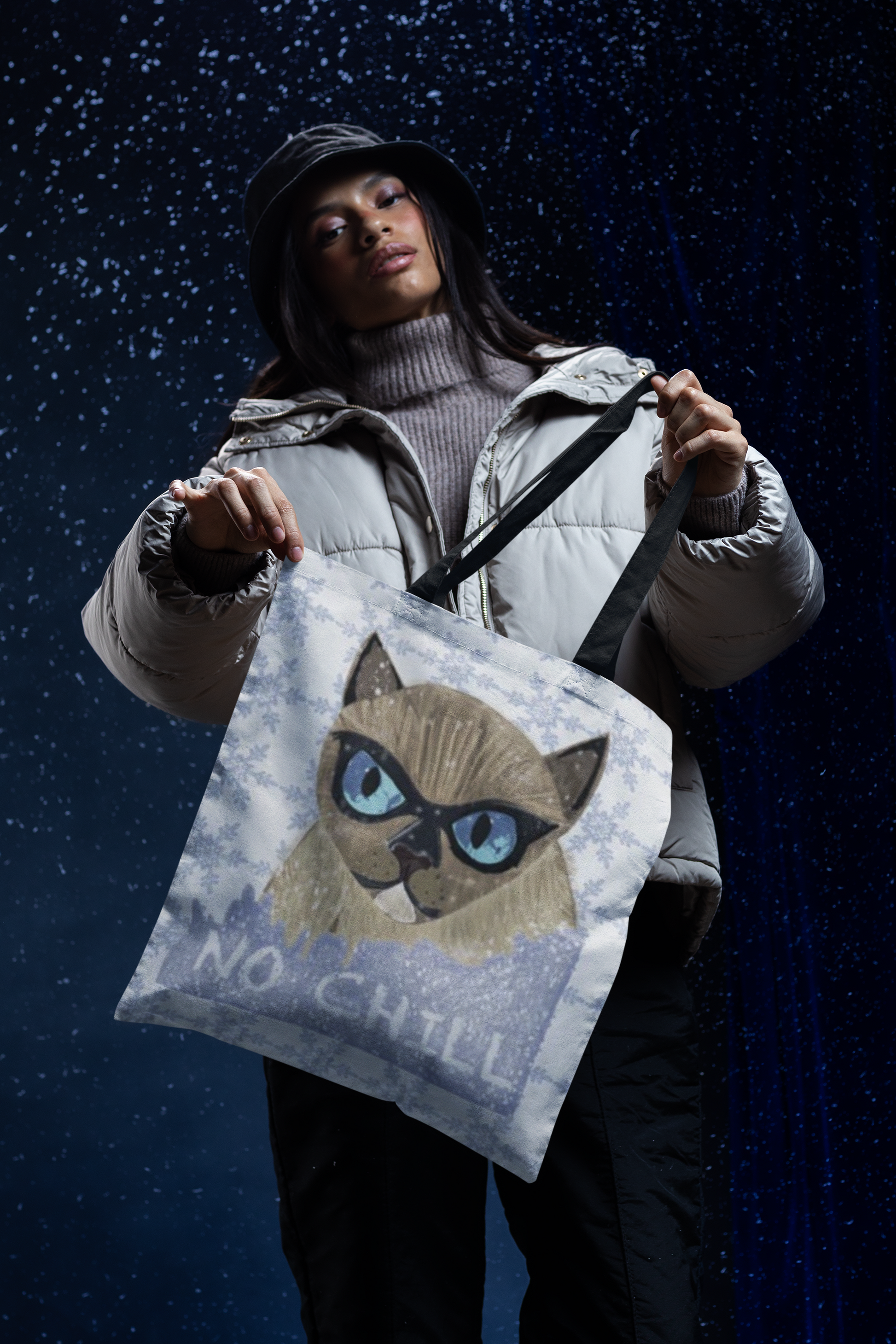 Woman in winter jacket and hat holding up the No Chill Cat snowflake tote bag against a dark snowy background