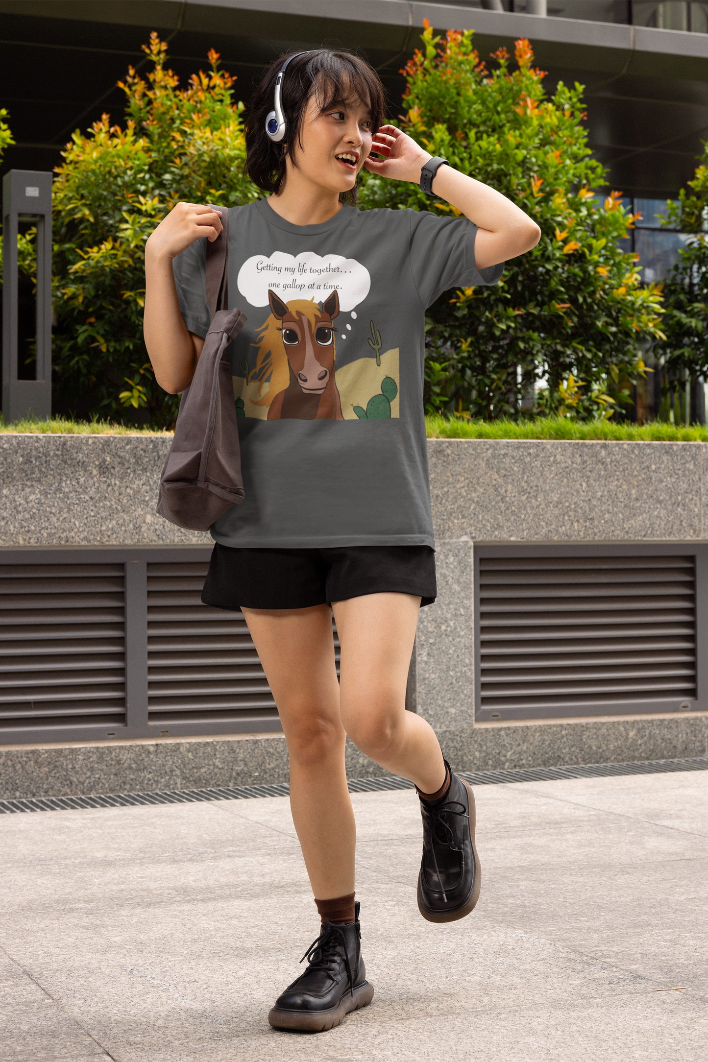 Woman wearing headphones and dark heather One Gallop at a Time t-shirt walking outdoors