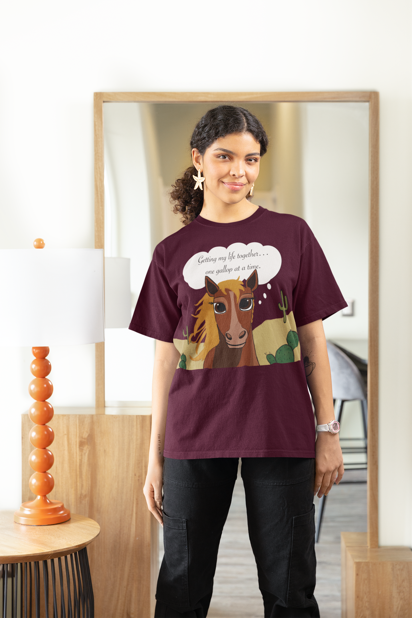 Woman with curly hair wearing maroon One Gallop at a Time t-shirt standing indoors