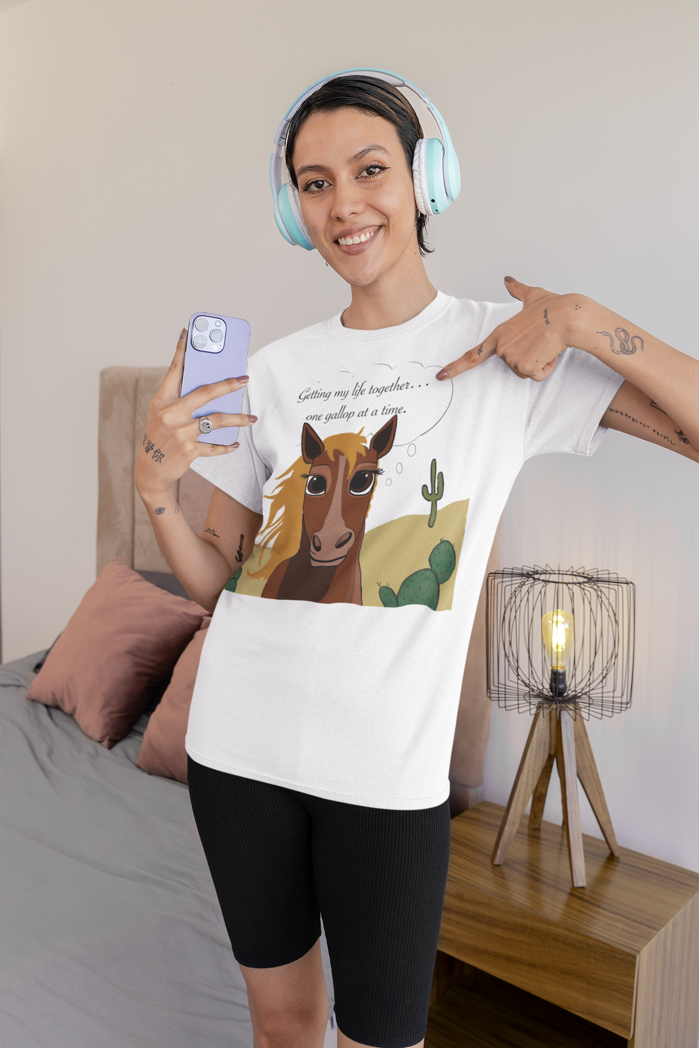 Smiling woman wearing white One Gallop at a Time t-shirt and headphones in bedroom setting