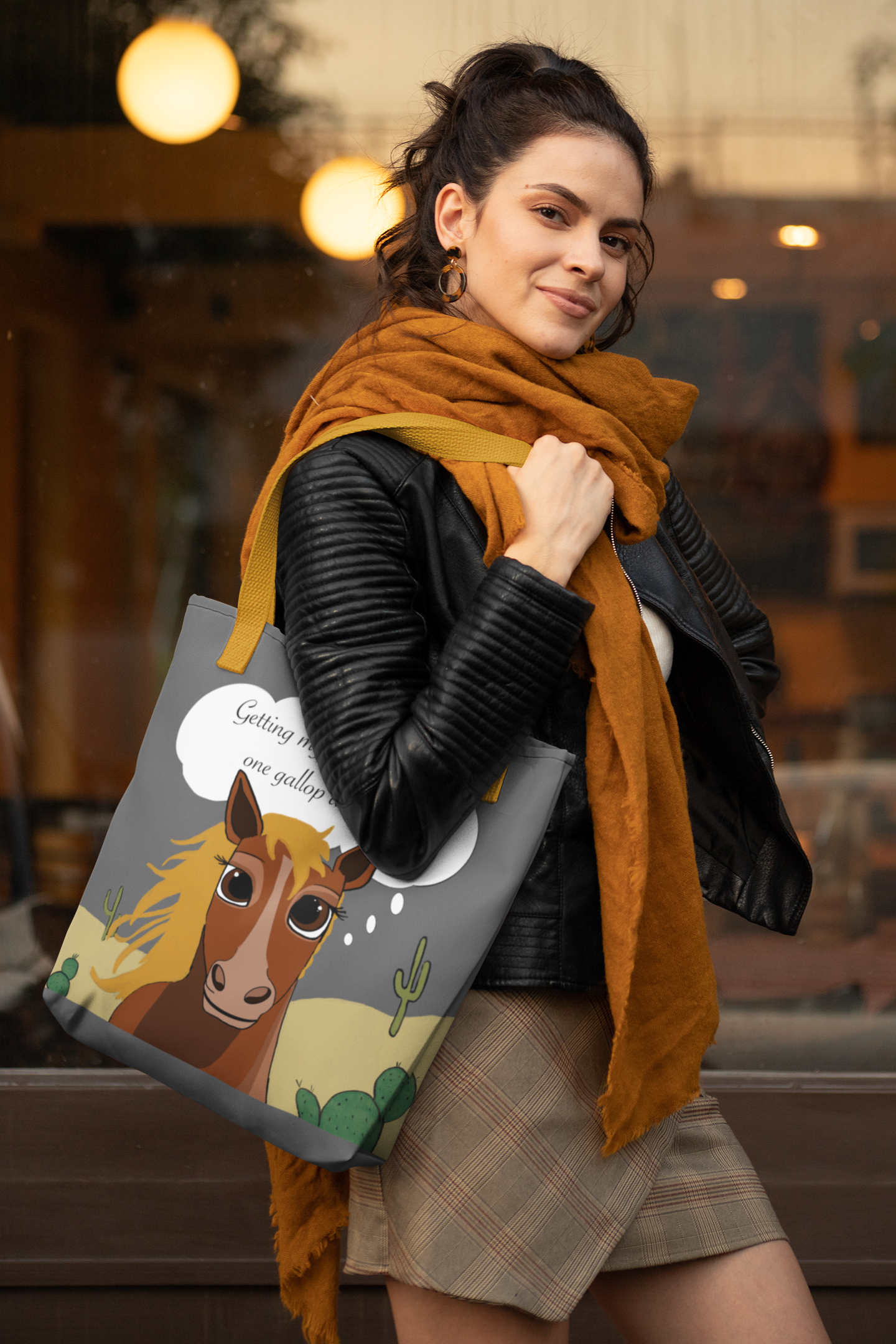 Woman in black leather jacket and orange scarf carrying One Gallop at a Time tote bag with cartoon horse design outside a storefront