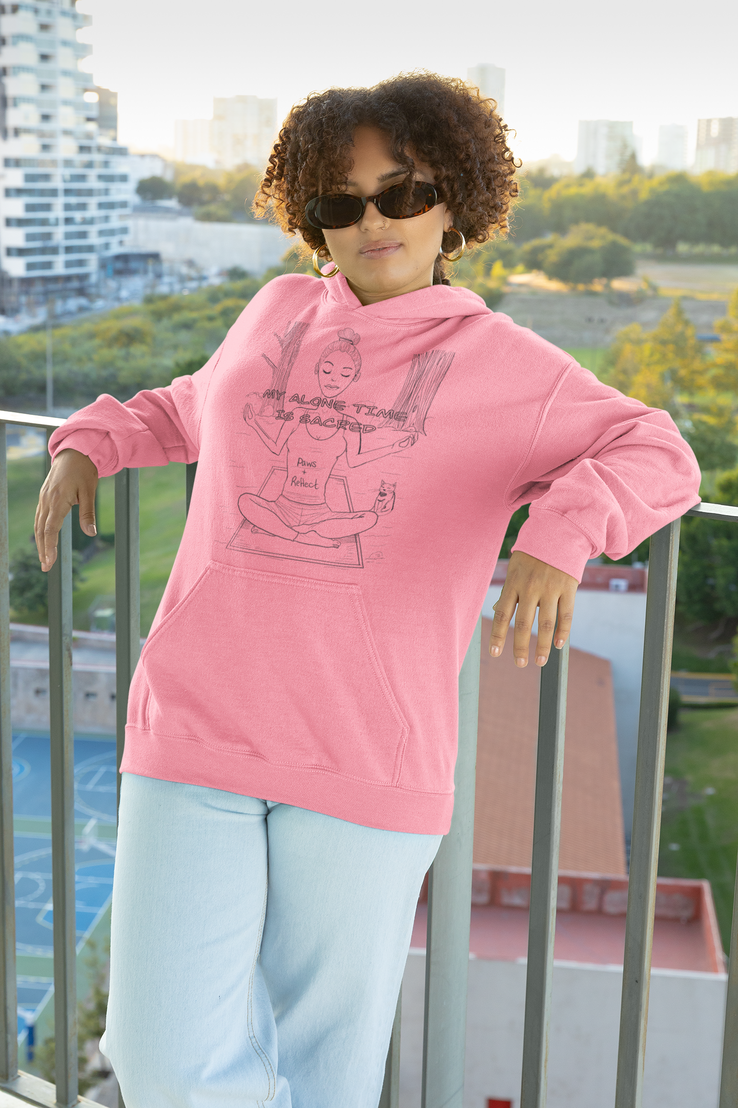 Woman with curly hair wearing pink Azalea Paws & Reflect hoodie with "My Alone Time is Sacred" design, leaning on balcony railing with city view