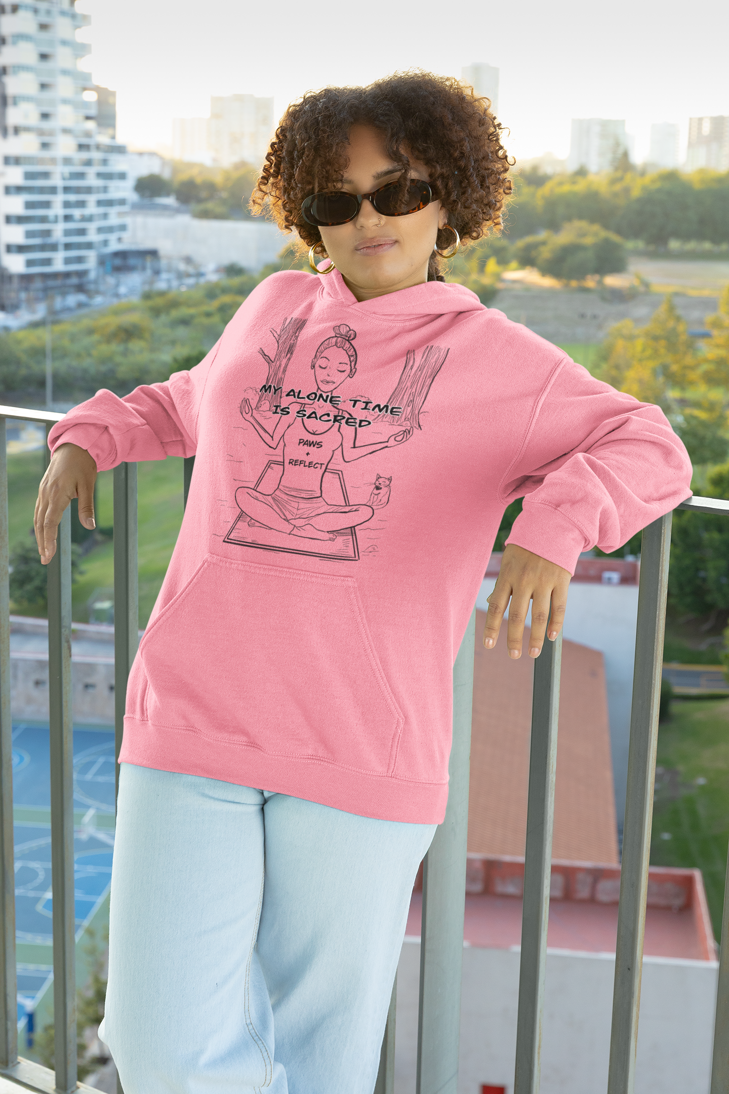 Woman with curly hair and sunglasses wearing an azalea pink Gildan hoodie featuring the darker “My Alone Time is Sacred” Paws & Reflect line art design, posing on a sunny balcony