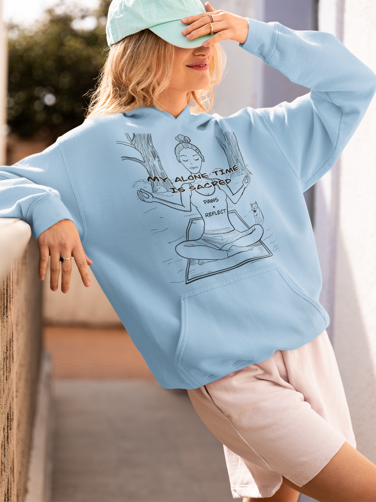 Smiling blonde woman in a light blue Gildan hoodie with the darker Paws & Reflect “My Alone Time is Sacred” design, posing on a balcony while adjusting her hat