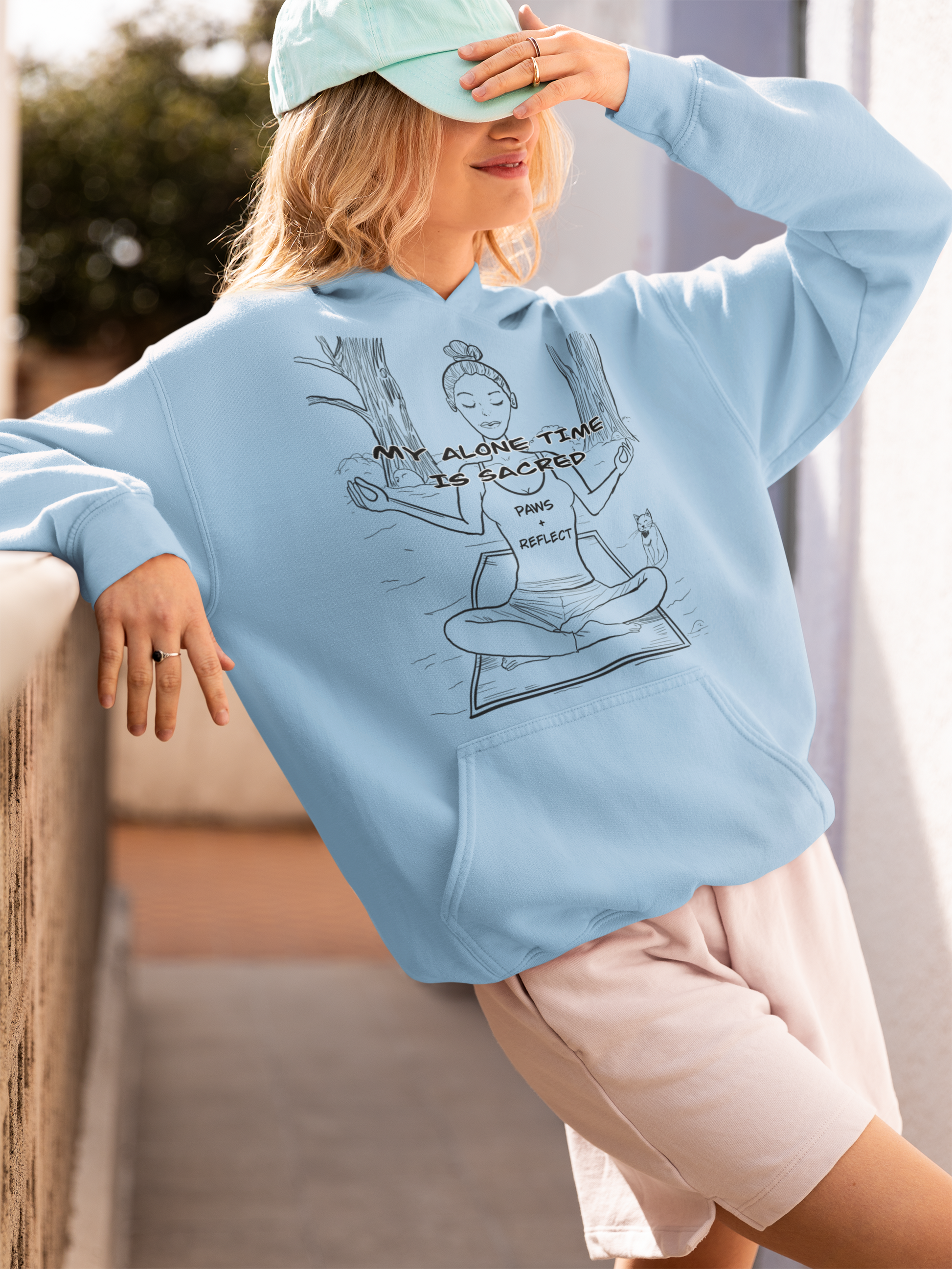 Smiling blonde woman in a light blue Gildan hoodie with the darker Paws & Reflect “My Alone Time is Sacred” design, posing on a balcony while adjusting her hat