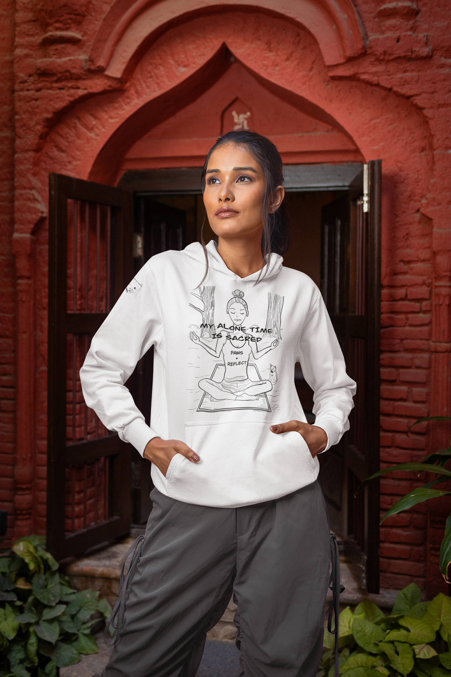 Woman standing in front of a red brick building wearing a white Gildan hoodie featuring the bold black line art “My Alone Time is Sacred” Paws & Reflect design
