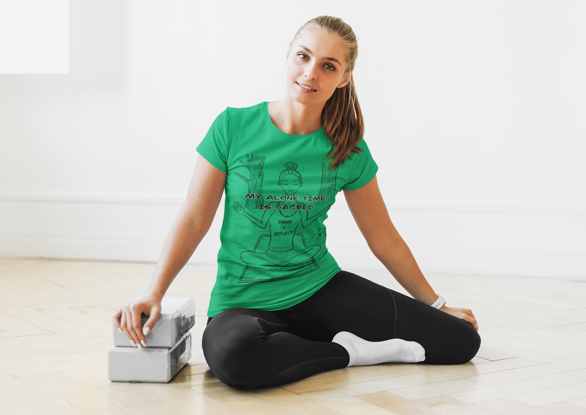Woman sitting on a wooden floor next to yoga blocks wearing an Irish green t-shirt featuring the bold “My Alone Time is Sacred” Paws & Reflect design