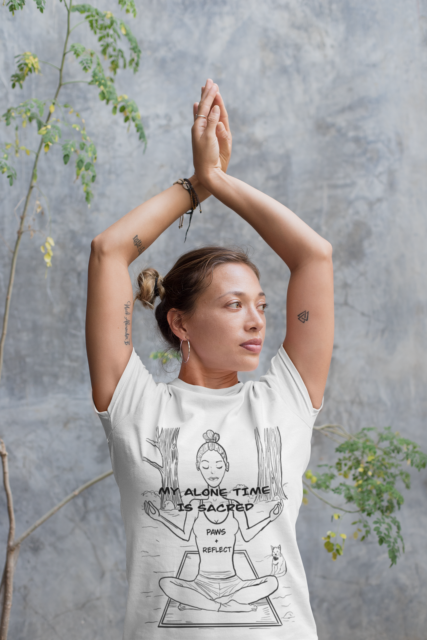 Yoga teacher with arms raised overhead in a prayer pose wearing a white t-shirt with the darker Paws & Reflect “My Alone Time is Sacred” design against a concrete wall with plants