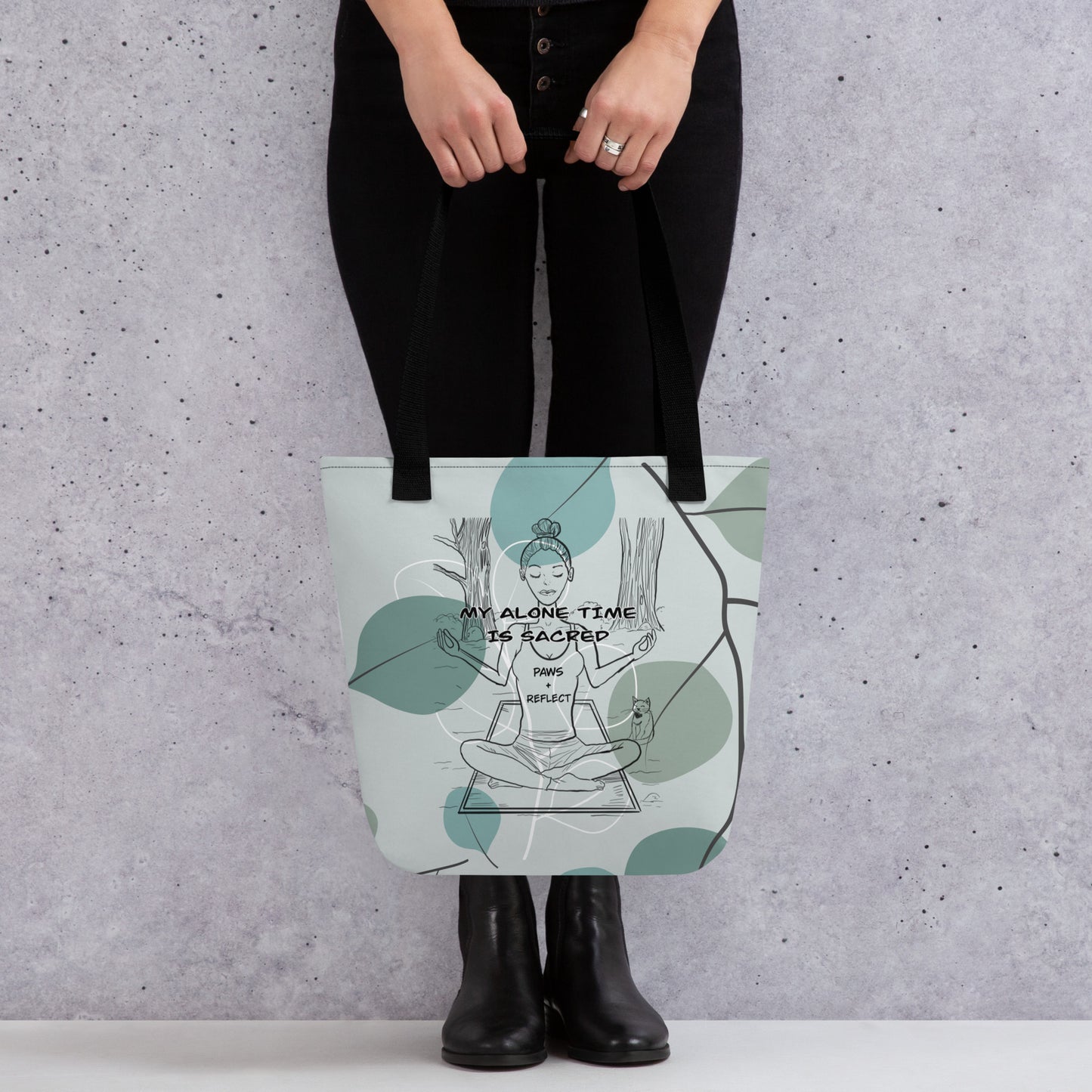 Back view of the sage green eucalyptus Paws & Reflect tote bag being held by a person in black pants and boots, clearly showing the meditating figure and cat design