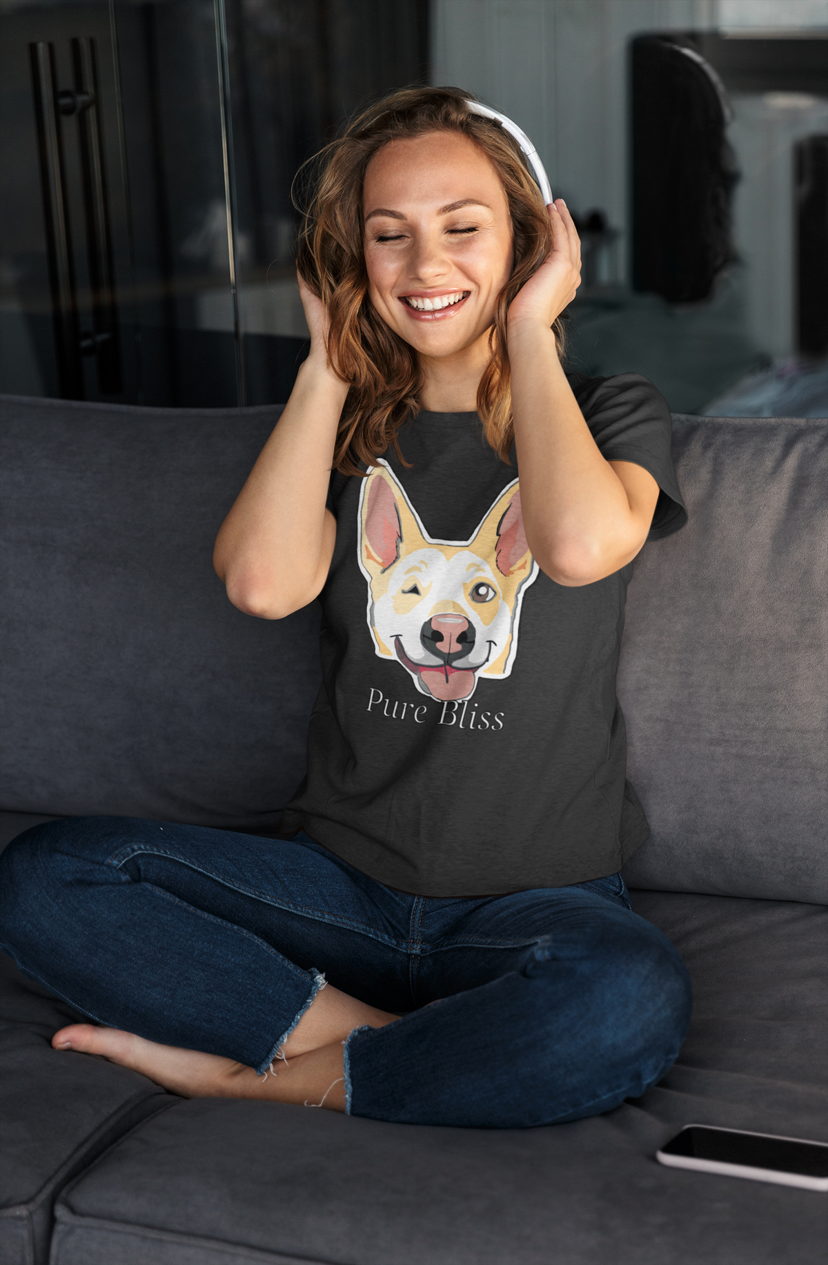 Woman sitting cross-legged on a gray couch in a black Pure Bliss t-shirt with headphones, smiling. Nico Inspired by GrungePup Gallery.