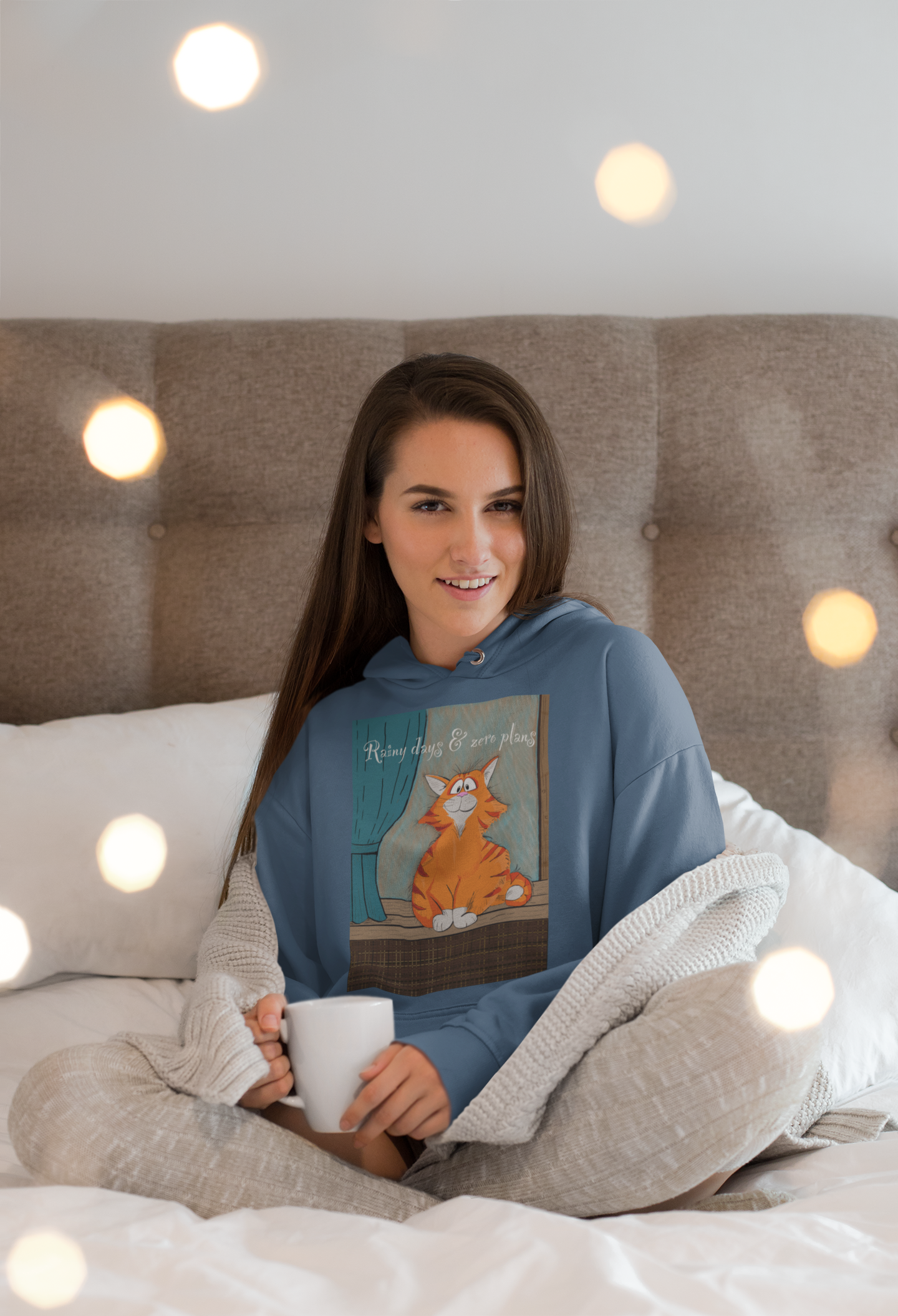 Woman sitting surrounded by twinkly lights wearing indigo blue Rainy Days & Zero Plans cat hoodie
