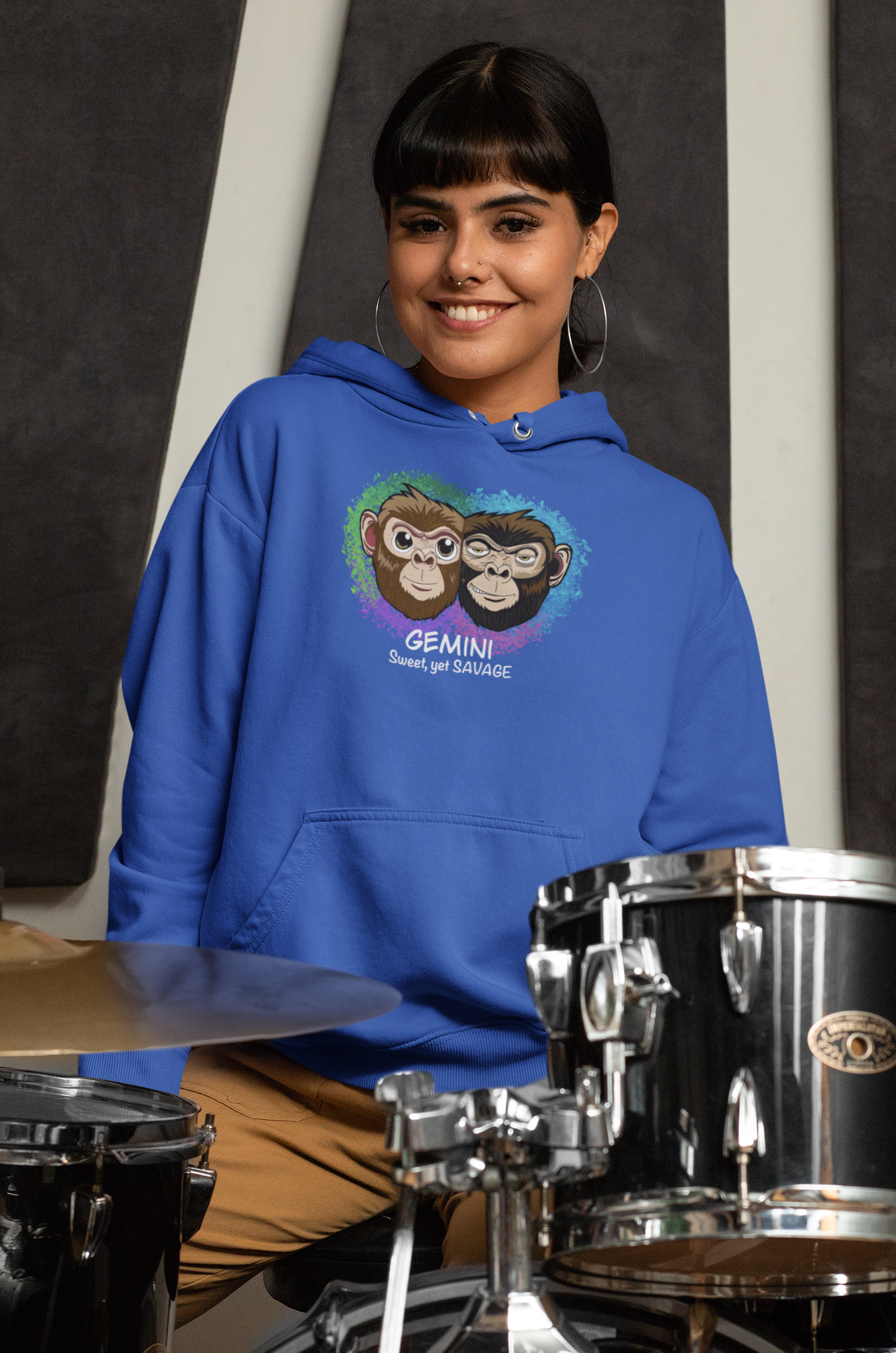 Smiling woman with dark hair and bangs wearing a team royal blue Gemini "Sweet, yet SAVAGE" hoodie sitting at a drum kit