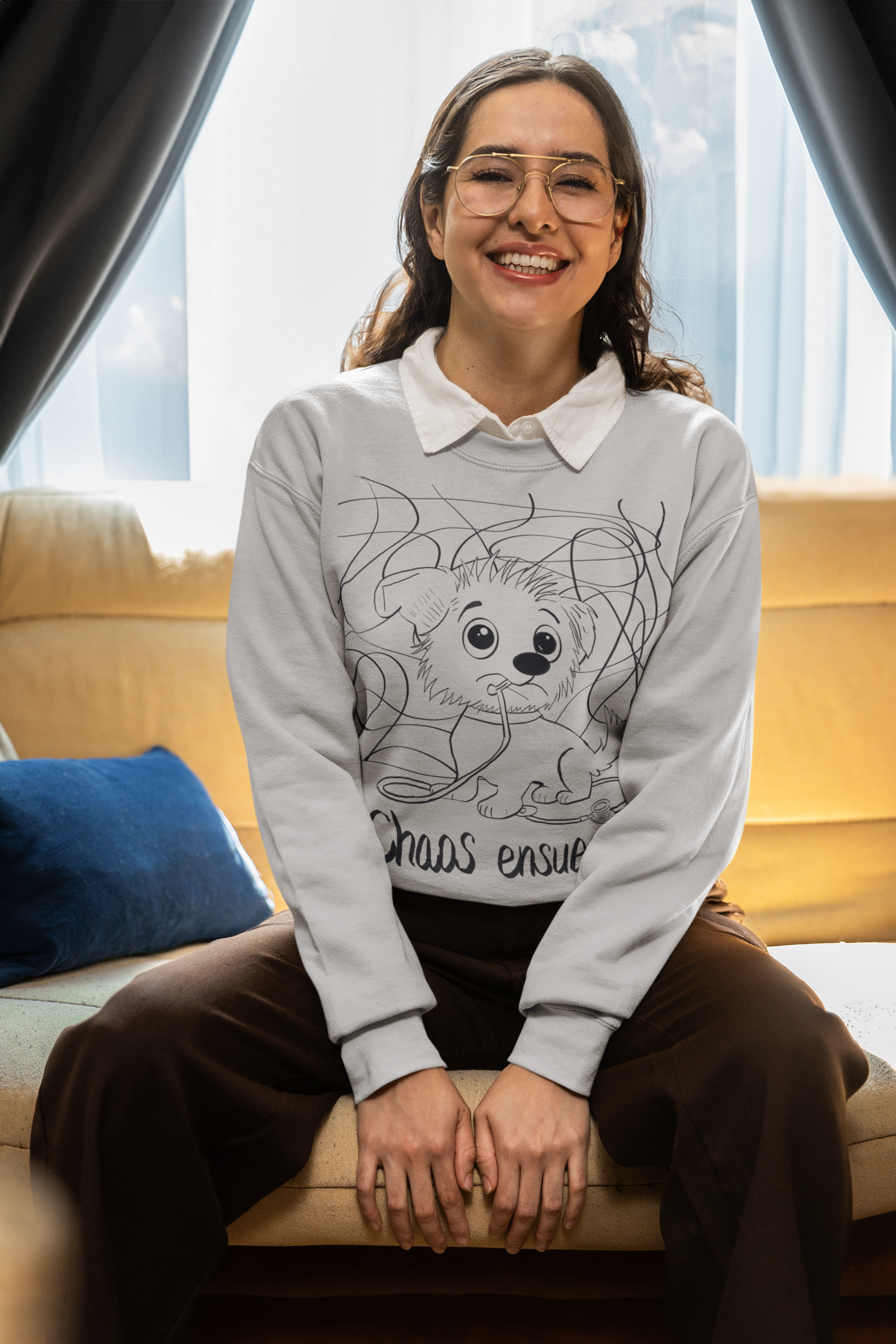 Happy woman with curly hair and gold glasses sitting and wearing a light grey Chaos Ensues sweatshirt