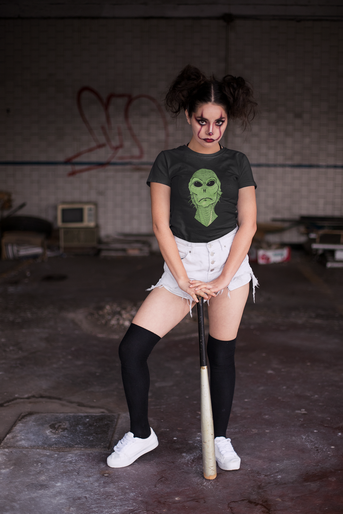 Woman with clown makeup holding a baseball bat in a warehouse wearing a black heather t-shirt with the Swamp Thang face