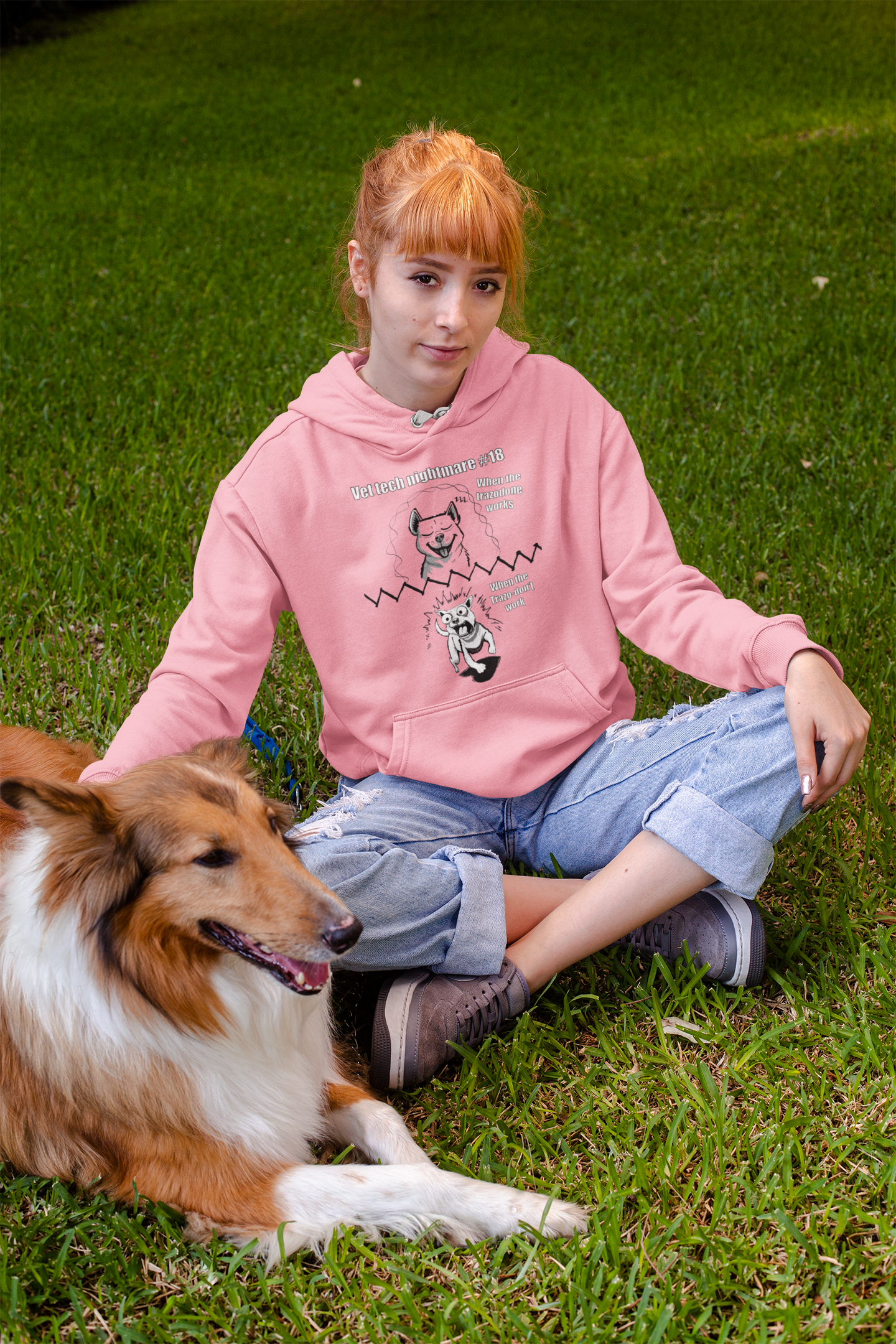 Woman with reddish hair sitting on grass wearing an azalea pink Gildan 18500 hoodie featuring Vet Tech Nightmare #18 with the happy relaxed dog on top and the demonic chaotic dog on bottom.