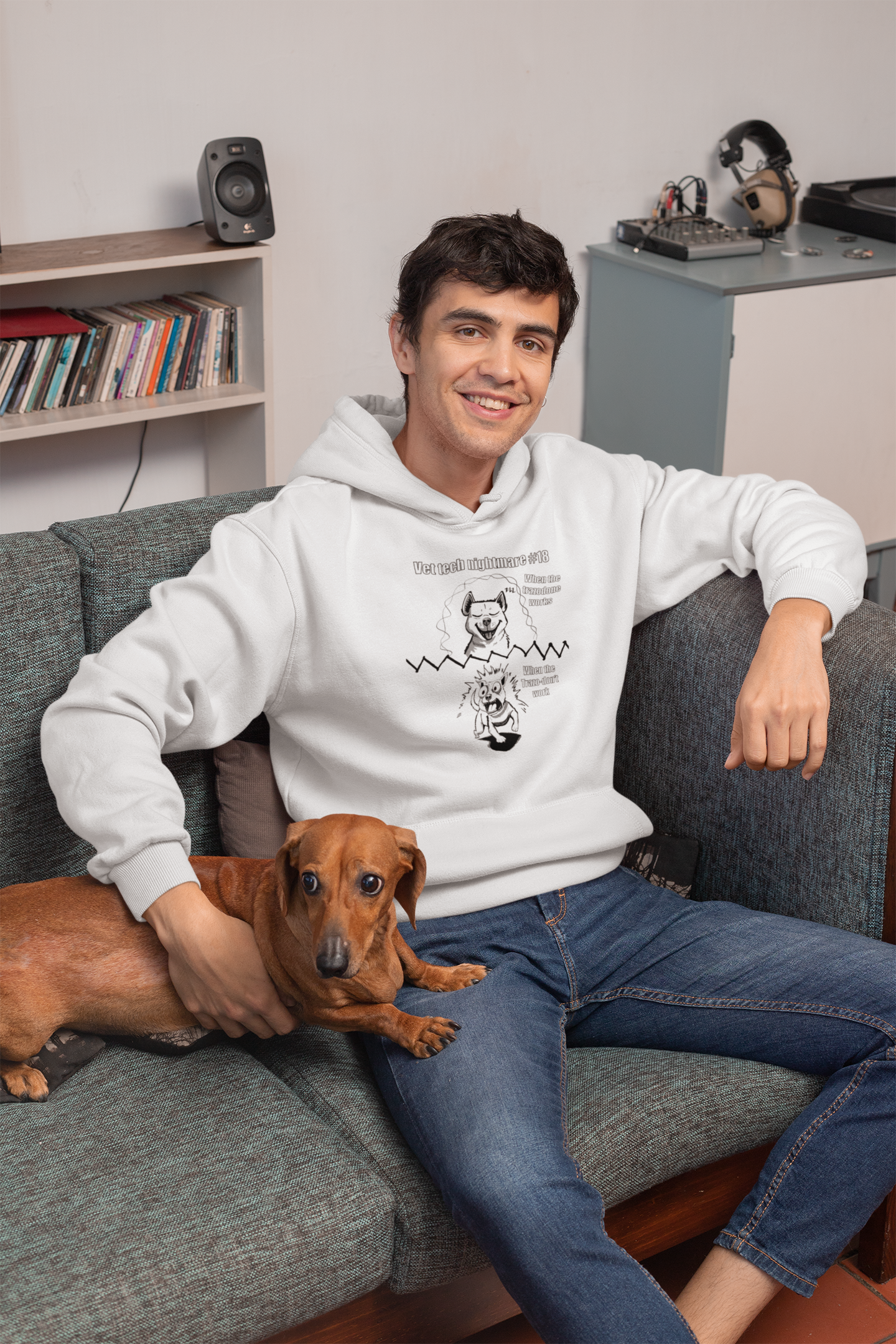 Man sitting on a couch holding a small dog while wearing a white Gildan 18500 hoodie displaying Vet Tech Nightmare #18 with the "When the Trazodone Works" and "When the Trazo-Don't Work" panels.