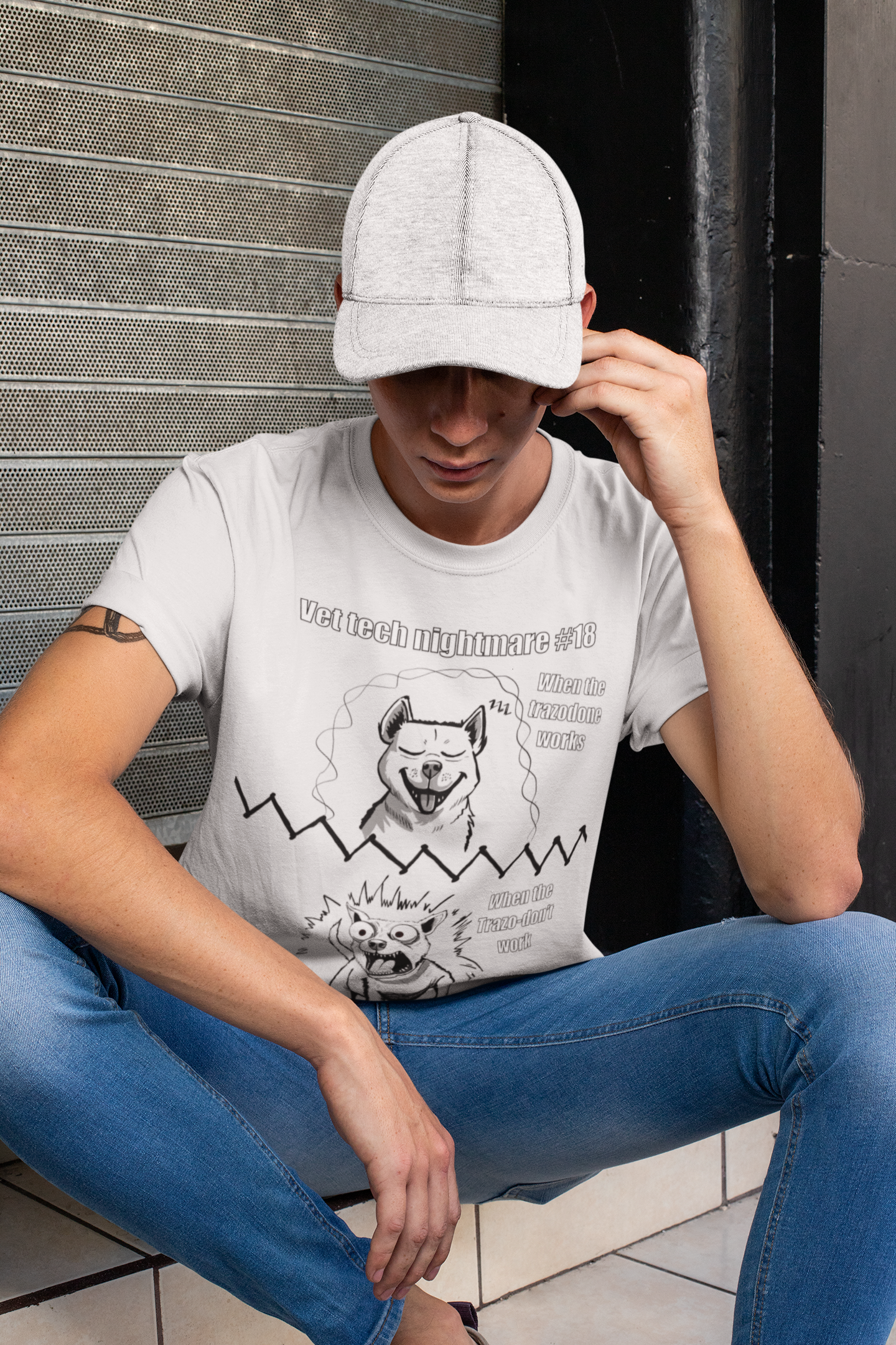 Young man wearing a grey ash Gildan 5000 t-shirt and a hat, sitting and looking at the camera, featuring Vet Tech Nightmare #18 with the split happy dog and chaotic demonic dog design.