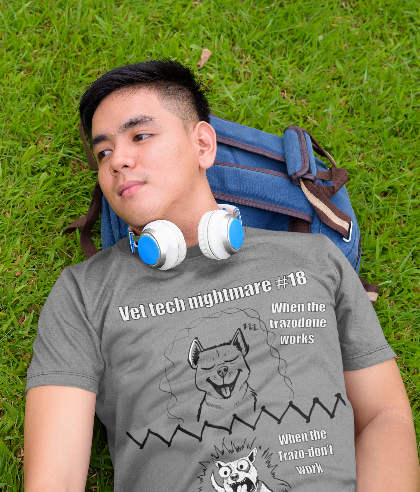 Young man laying on grass wearing a graphite heather Gildan 5000 t-shirt with Vet Tech Nightmare #18 design showing the relaxed happy dog on top and the wild demonic dog on bottom.