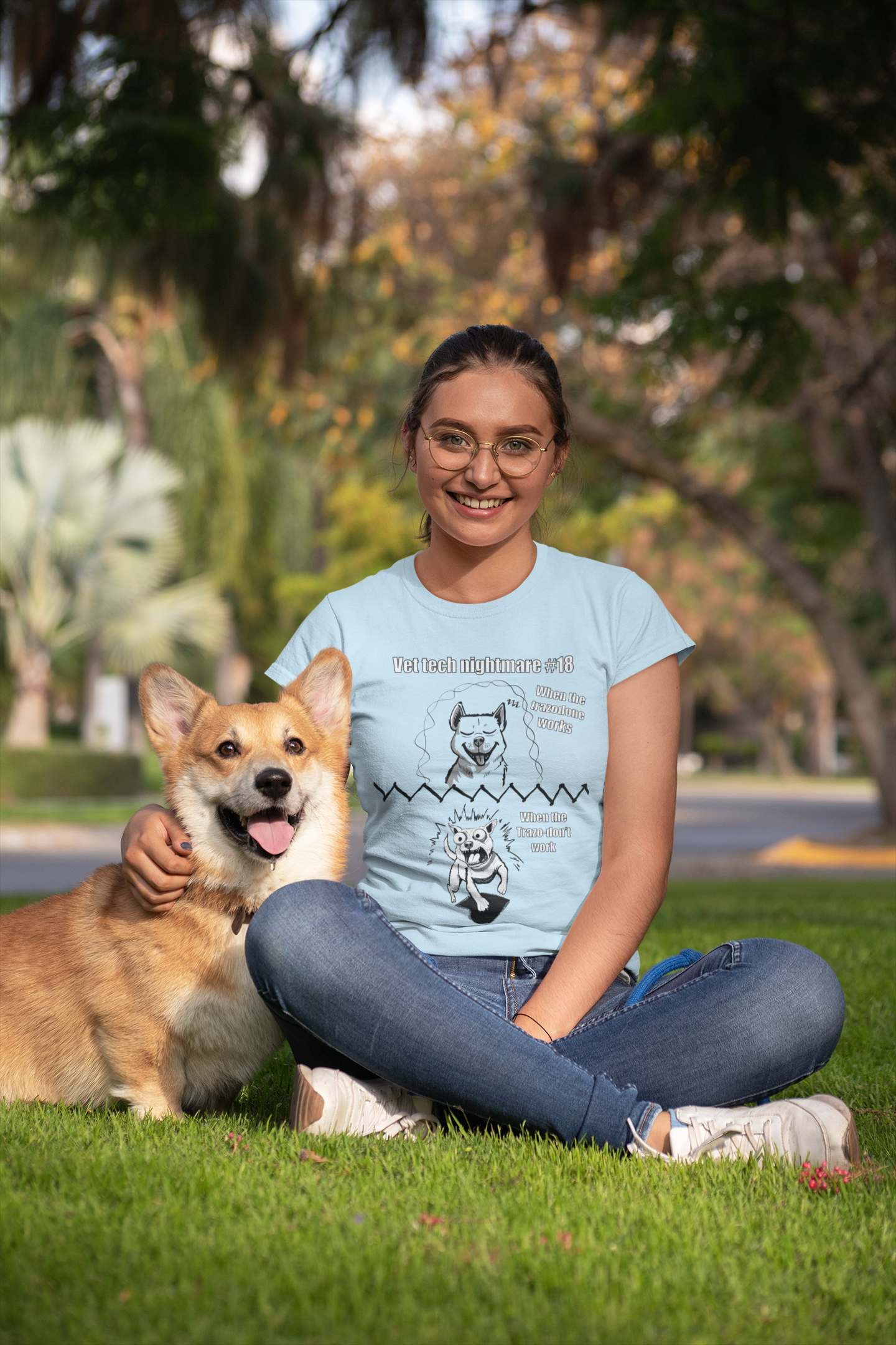 Smiling woman sitting on grass next to a corgi wearing a light blue Gildan 5000 t-shirt with the Vet Tech Nightmare #18 design showing the "When the Trazodone Works" happy dog and "When the Trazo-Don't Work" demonic dog.