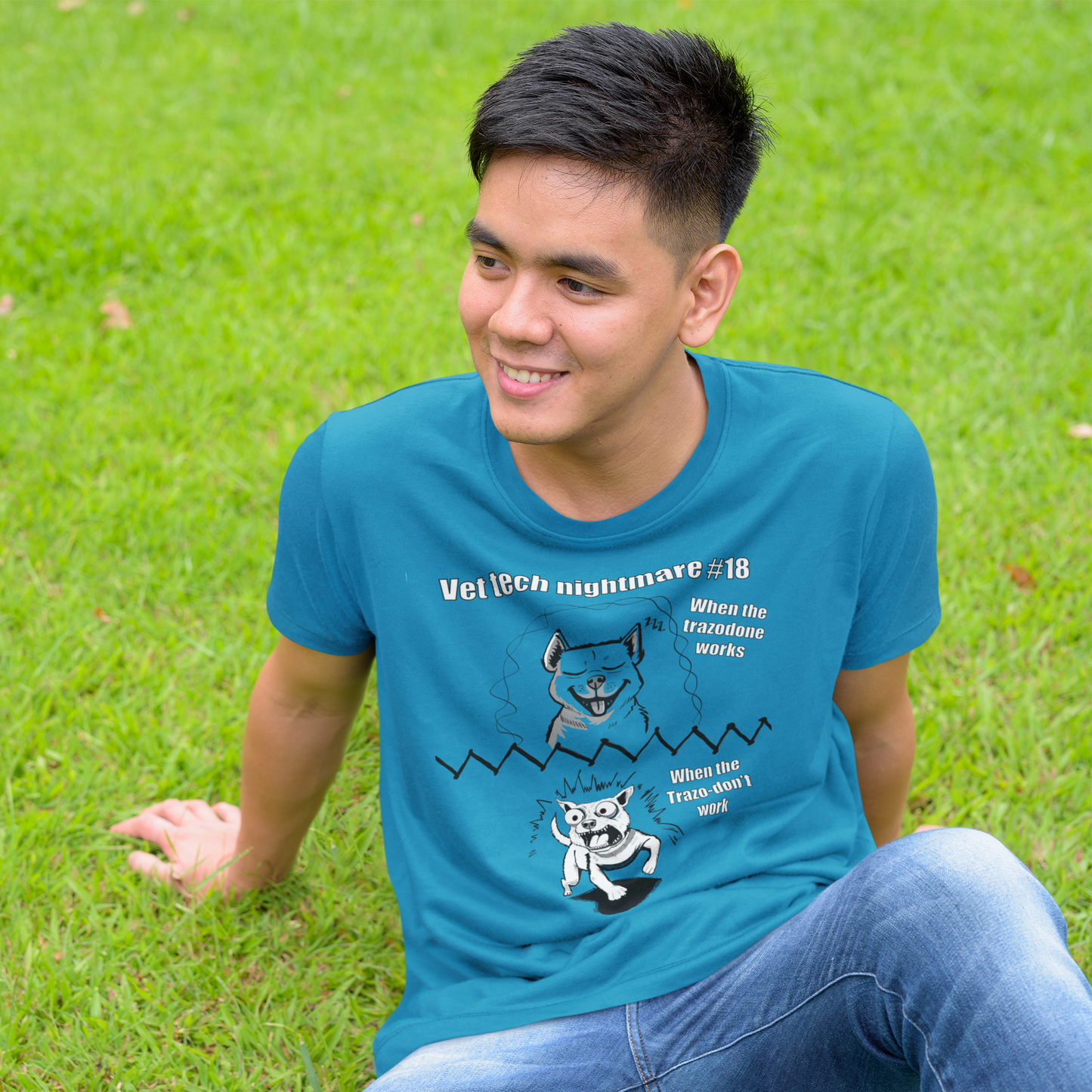 Young man with short black hair sitting on grass wearing a sapphire blue Gildan 5000 t-shirt featuring Vet Tech Nightmare #18 with the happy dog ("When the Trazodone Works") on top and the demonic chaotic dog ("When the Trazo-Don't Work") on bottom.