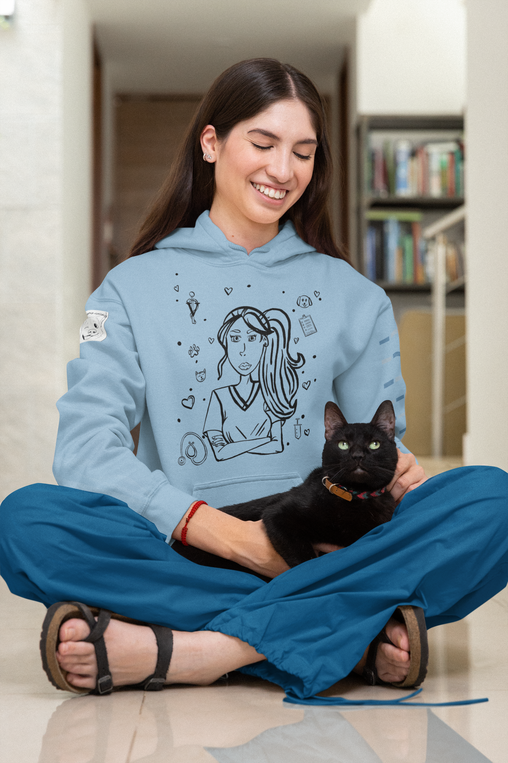 Woman with long dark hair sitting cross-legged on the floor, smiling with eyes closed while holding a black cat, wearing a light blue Vet Vibe hoodie with the darkened line-art vet girl design.