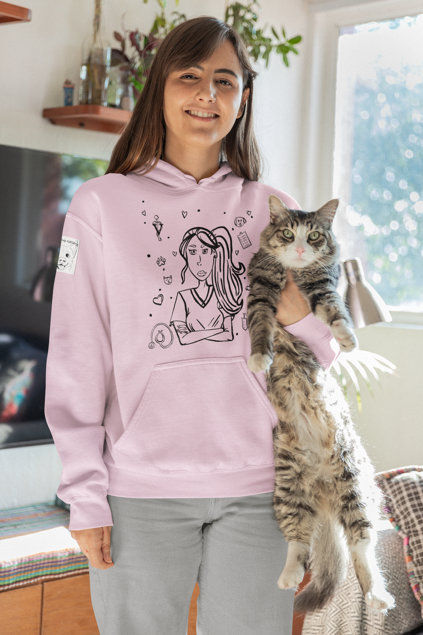 Woman with long brown hair smiling while holding a tabby cat and wearing a light pink Vet Vibe hoodie featuring the darkened line-art vet girl design with hearts, paw prints, and medical icons.