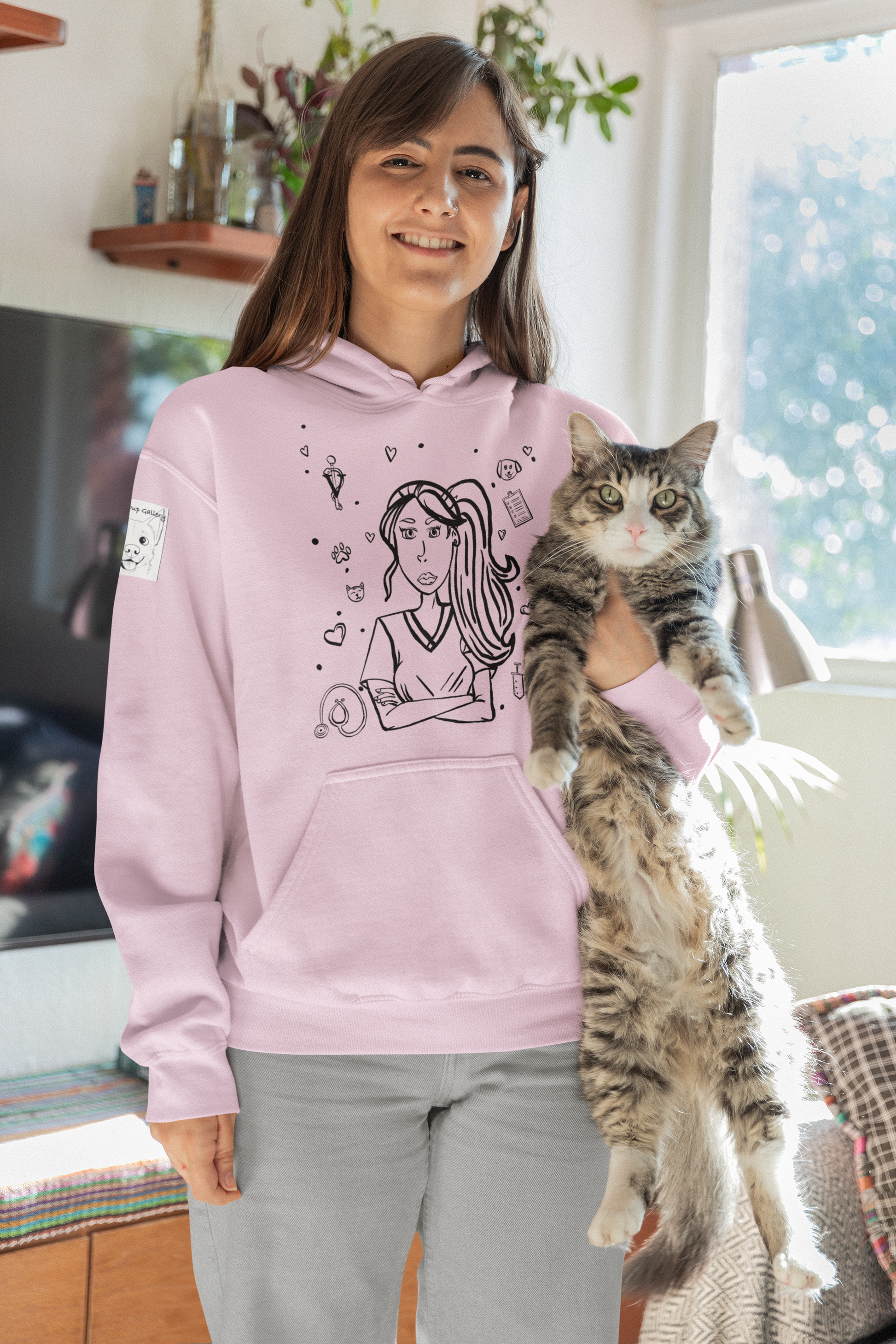 Woman with long brown hair smiling while holding a tabby cat and wearing a light pink Vet Vibe hoodie featuring the darkened line-art vet girl design with hearts, paw prints, and medical icons.