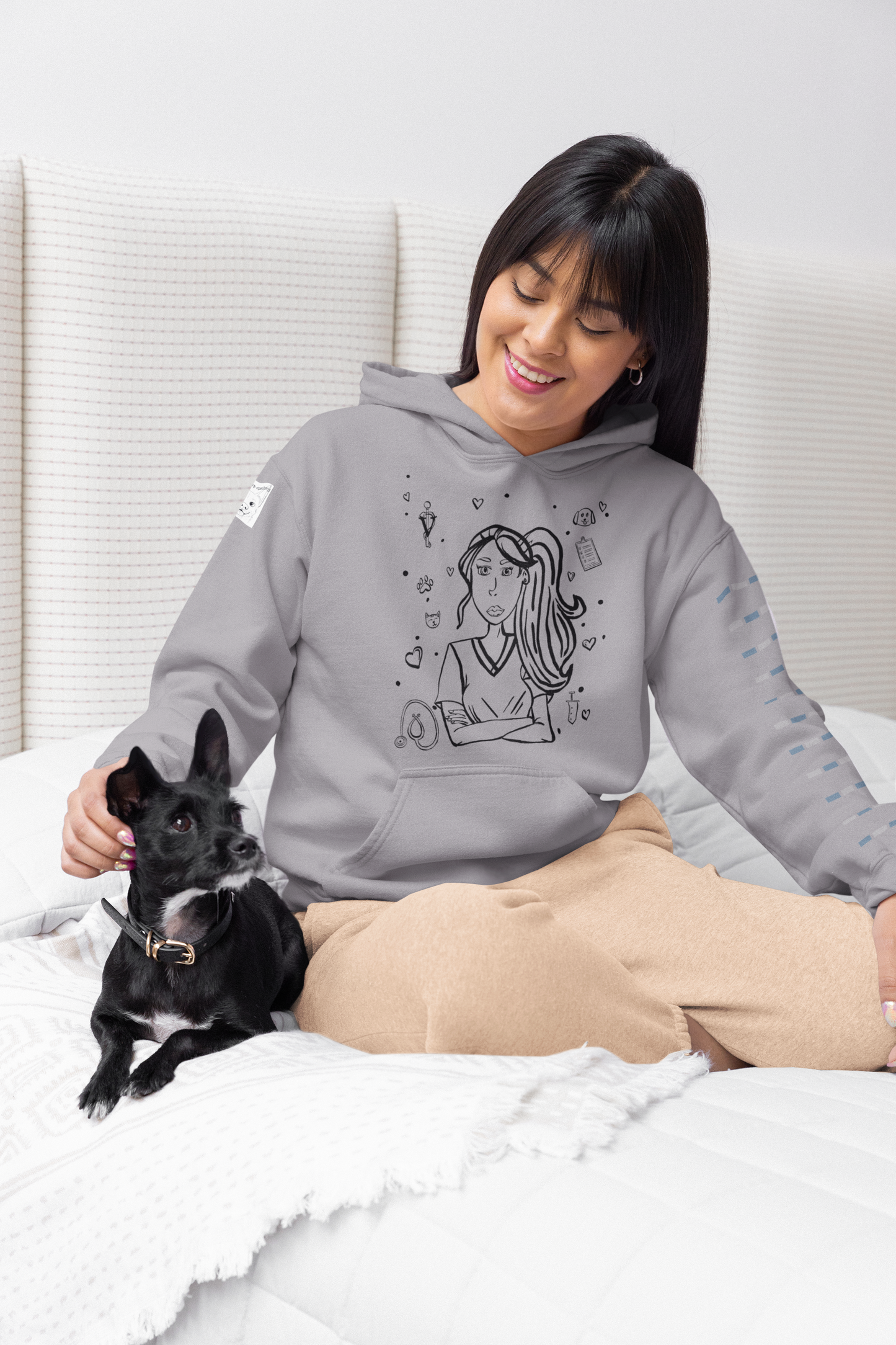 Woman with dark hair sitting on a bed and smiling while petting a small black dog, wearing a sport grey Vet Vibe hoodie with the darkened line-art vet girl design.