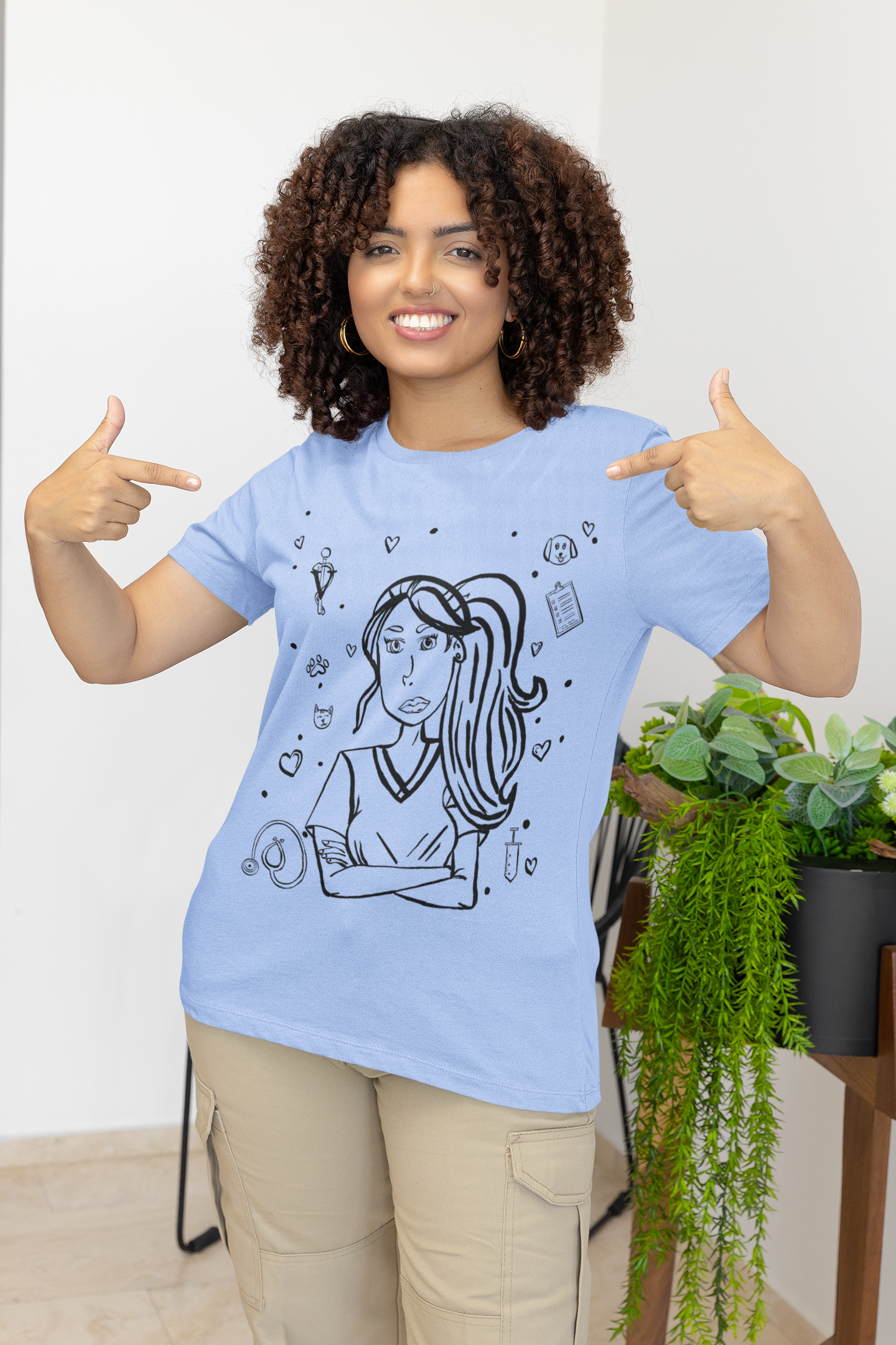 Woman with curly hair smiling and pointing at the design on her heather carolina blue Vet Vibe t-shirt featuring the darkened line-art vet girl with crossed arms.