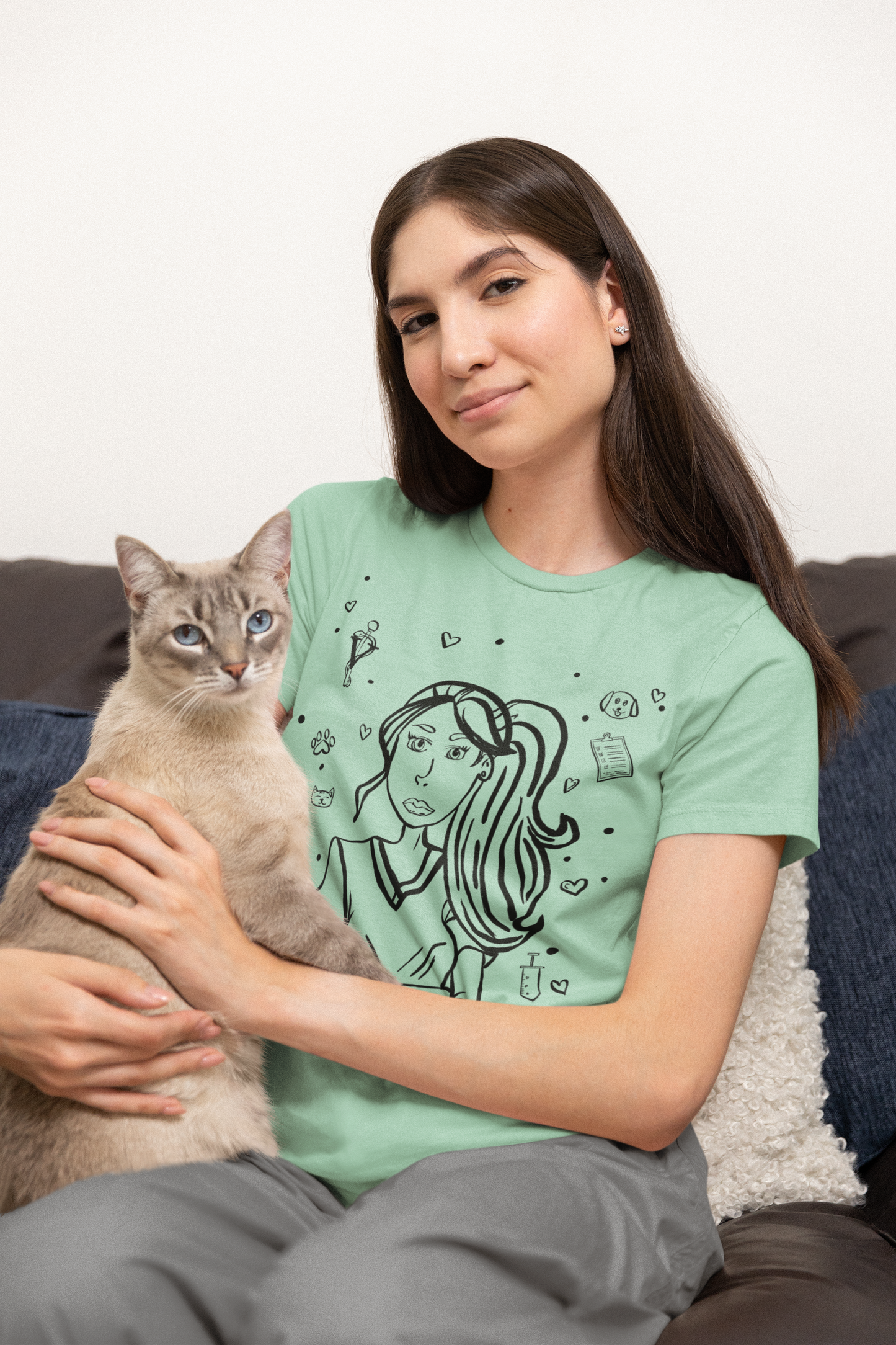 Woman with long brown hair smiling while holding a light-colored cat and wearing a heather prism mint Vet Vibe t-shirt featuring the darkened line-art vet girl design.