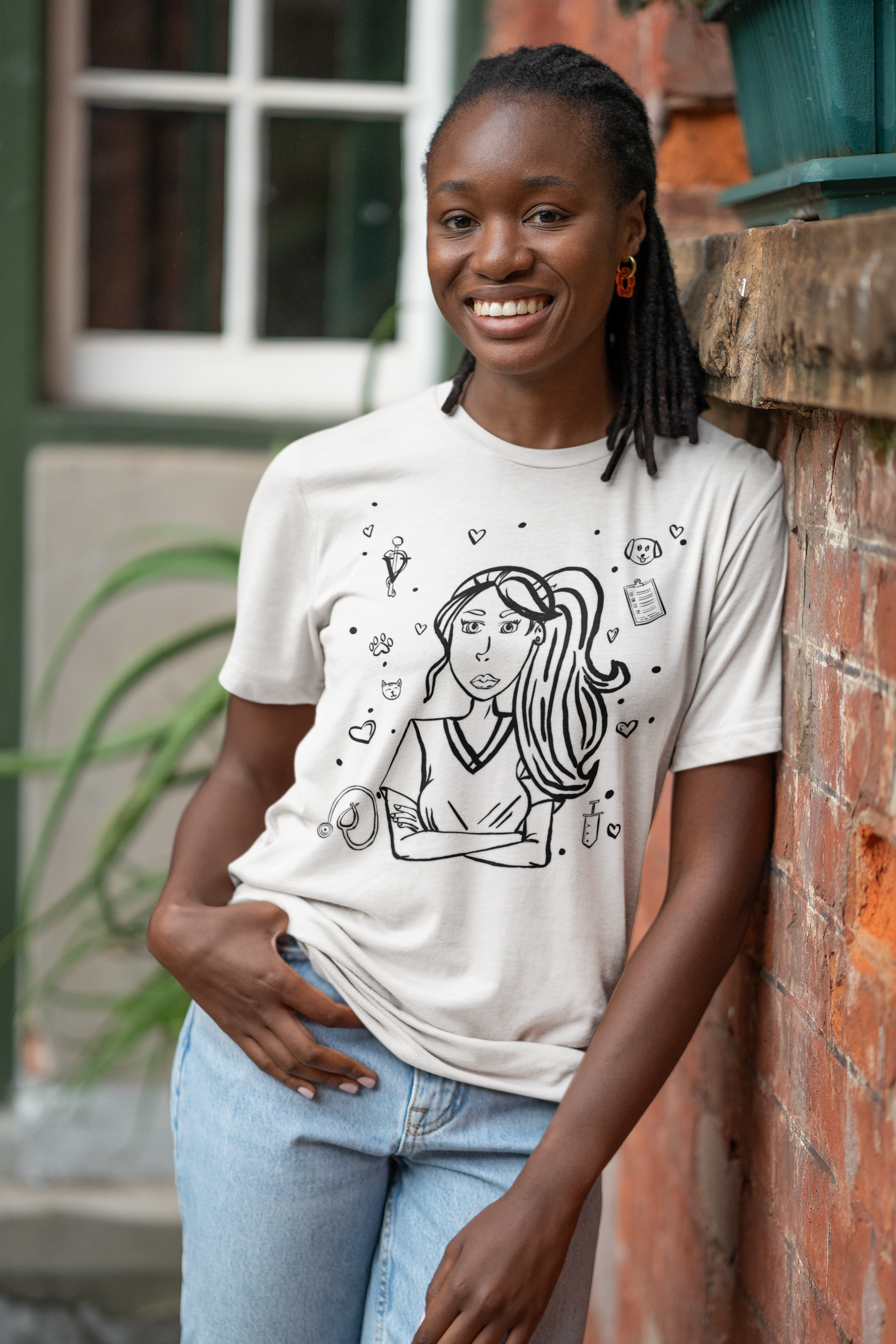 Black woman with braided hair smiling and leaning against a red brick wall while wearing a white Vet Vibe t-shirt featuring the darkened line-art vet girl design surrounded by hearts, paw prints, and medical icons.