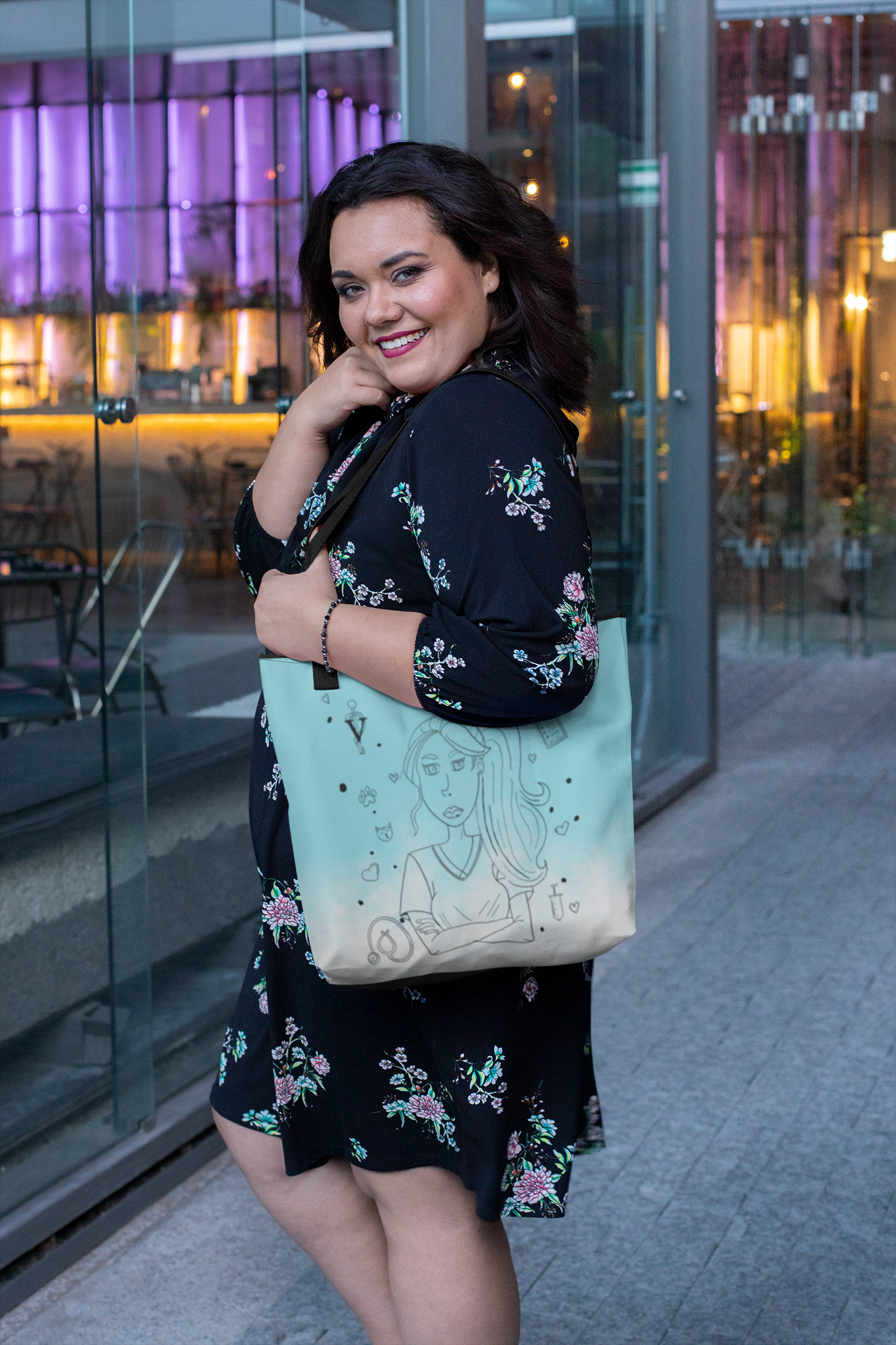Smiling woman with dark wavy hair wearing a black floral dress carrying the light blue Vet Vibe tote bag showing the front girl illustration design outdoors