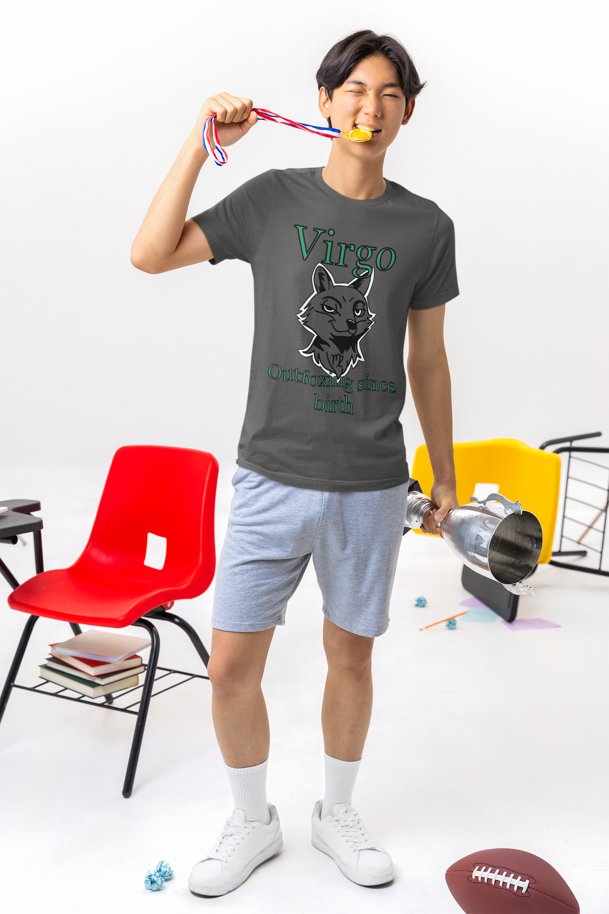 Young man wearing an asphalt Bella + Canvas Virgo Outfoxing Since Birth t-shirt in a classroom while holding a trophy and biting a medal.