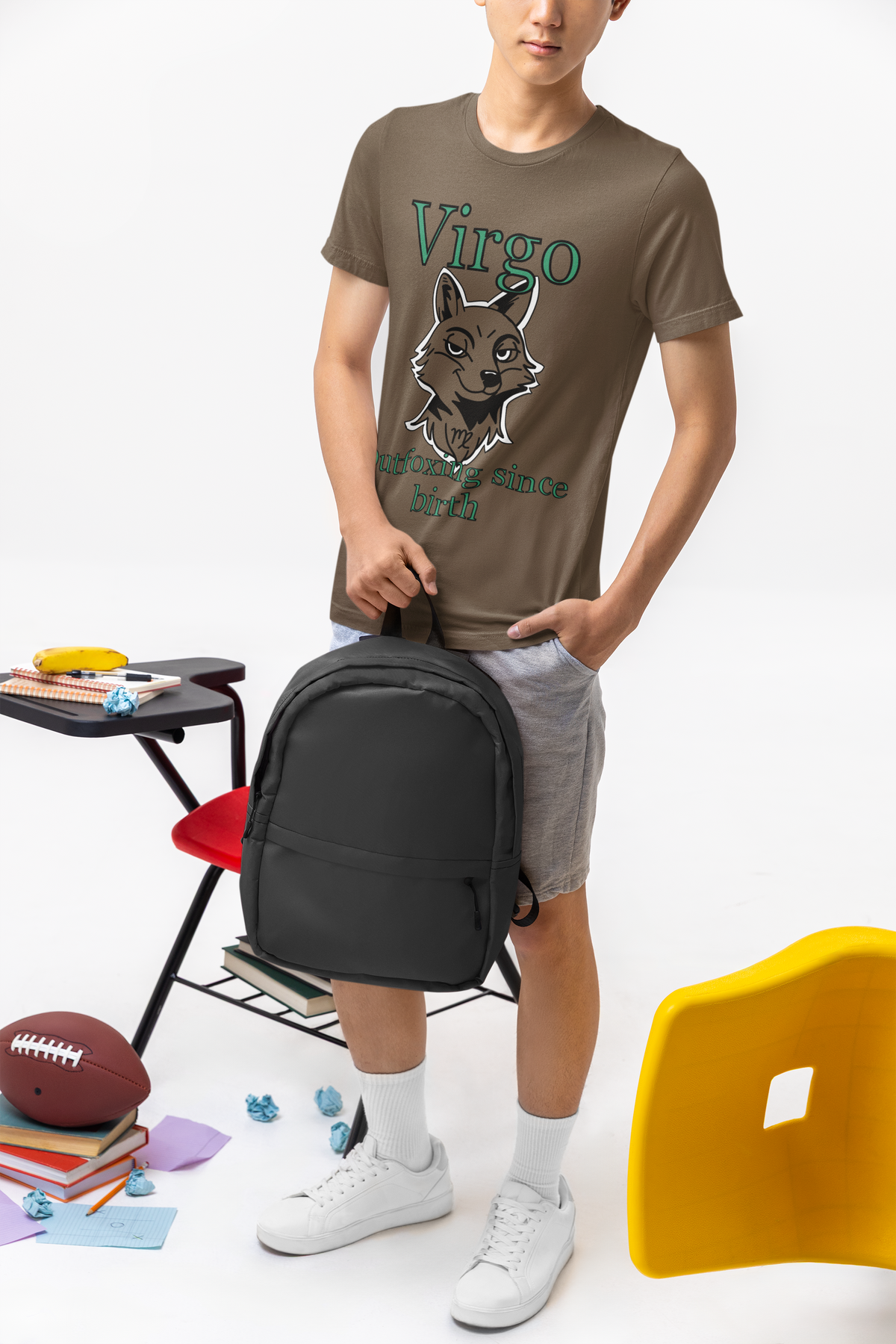 Young man wearing a heather brown Bella + Canvas Virgo Outfoxing Since Birth t-shirt while holding a black backpack in a classroom setting.