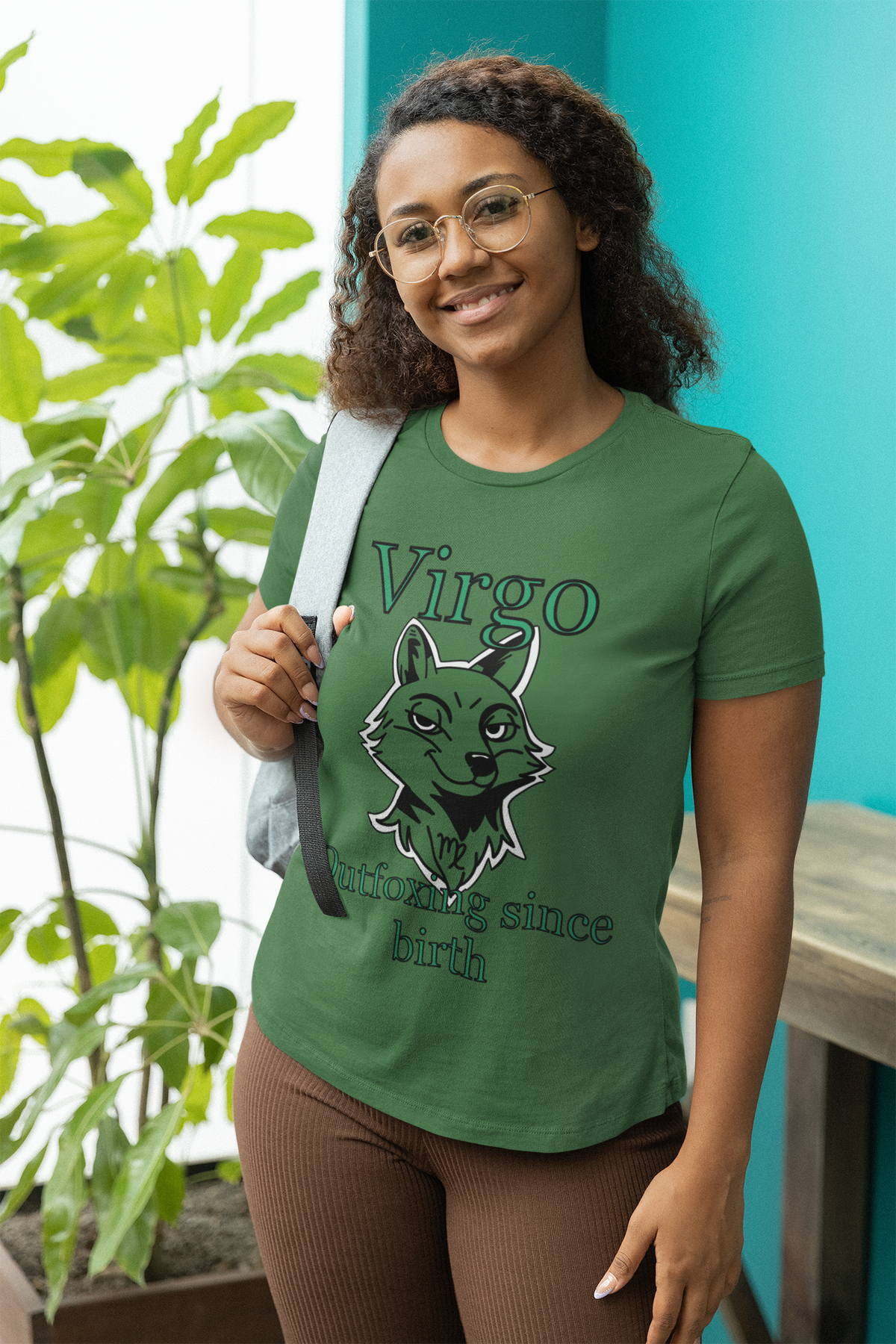 Woman with curly hair and glasses wearing a heather grass green Bella + Canvas Virgo Outfoxing Since Birth t-shirt while carrying a backpack.