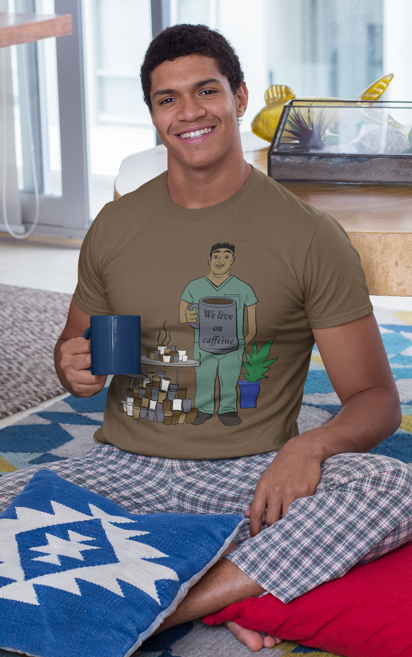 Young man sitting on floor wearing heather brown We Live on Caffeine t-shirt holding blue coffee mug – relatable vet tech burnout design by GrungePup Vet Chaos