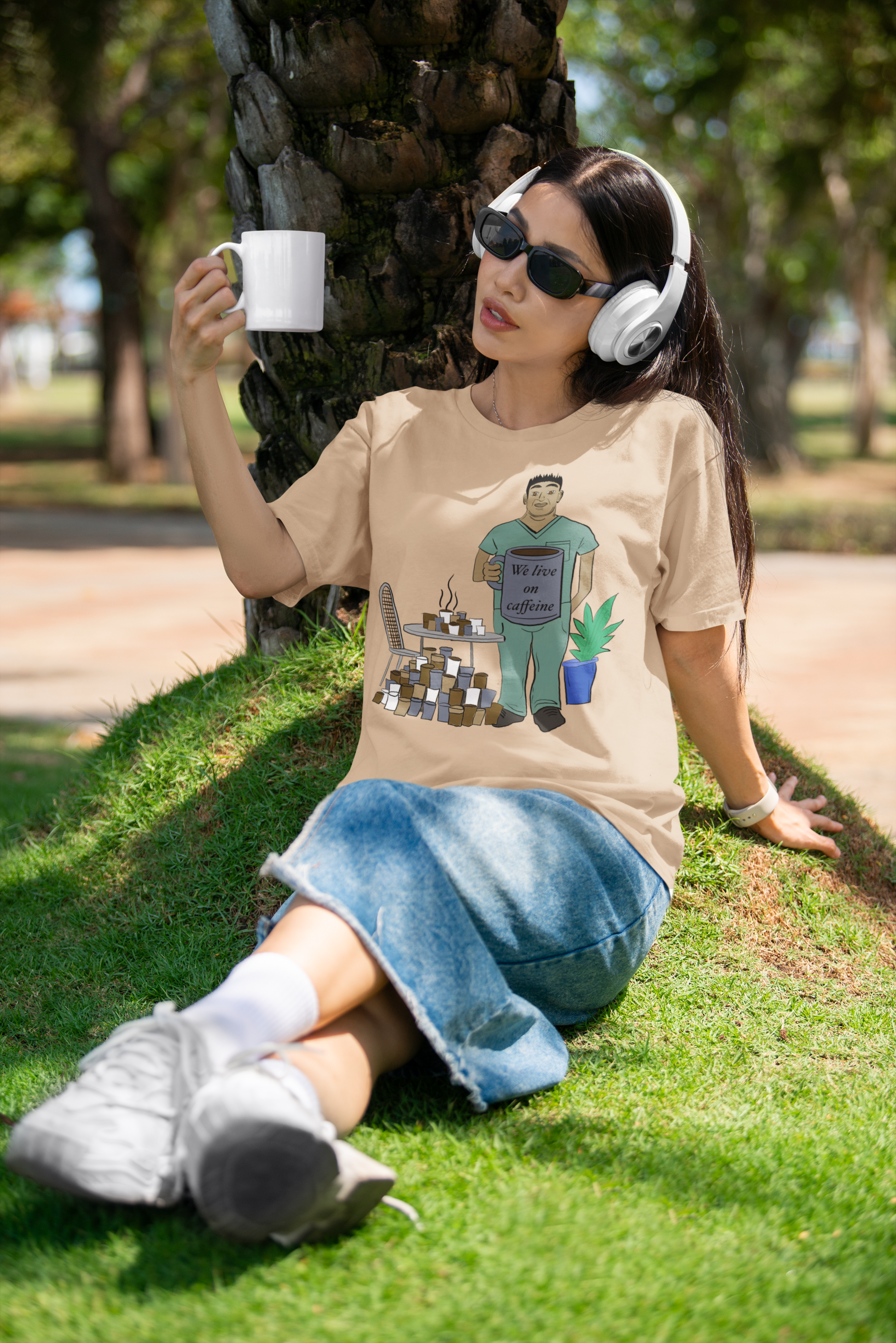 Woman sitting outside under tree wearing tan We Live on Caffeine t-shirt holding white coffee mug with headphones – GrungePup Vet Chaos vet tech burnout humor