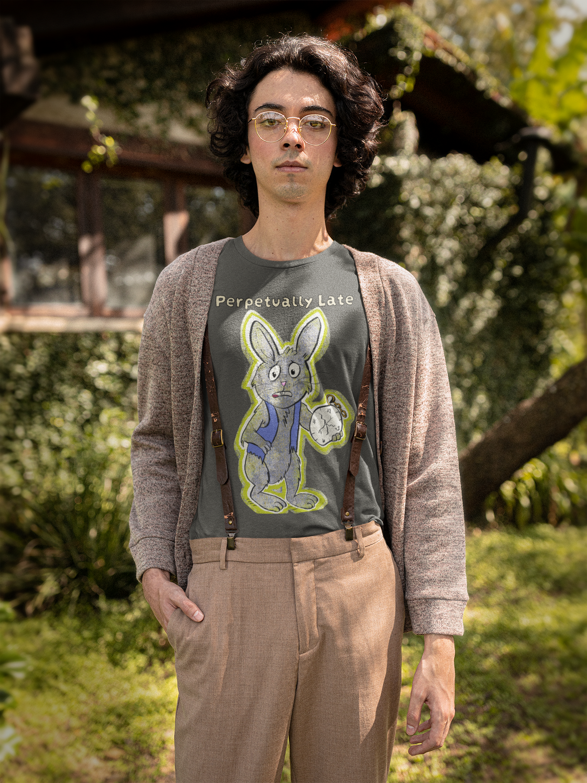 Man in gold glasses standing by a countryside house in an asphalt grey White Rabbit Perpetually Late t-shirt, cardigan, and suspenders. GrungePup That Girl, Alice.