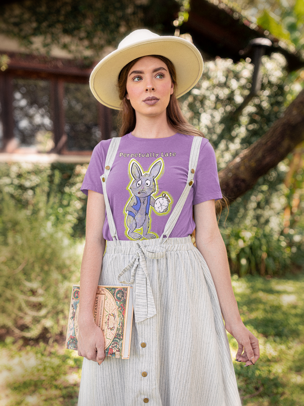 Woman in a wide cream hat holding a book, wearing a heather team purple White Rabbit Perpetually Late t-shirt with striped overalls outdoors. GrungePup That Girl, Alice.